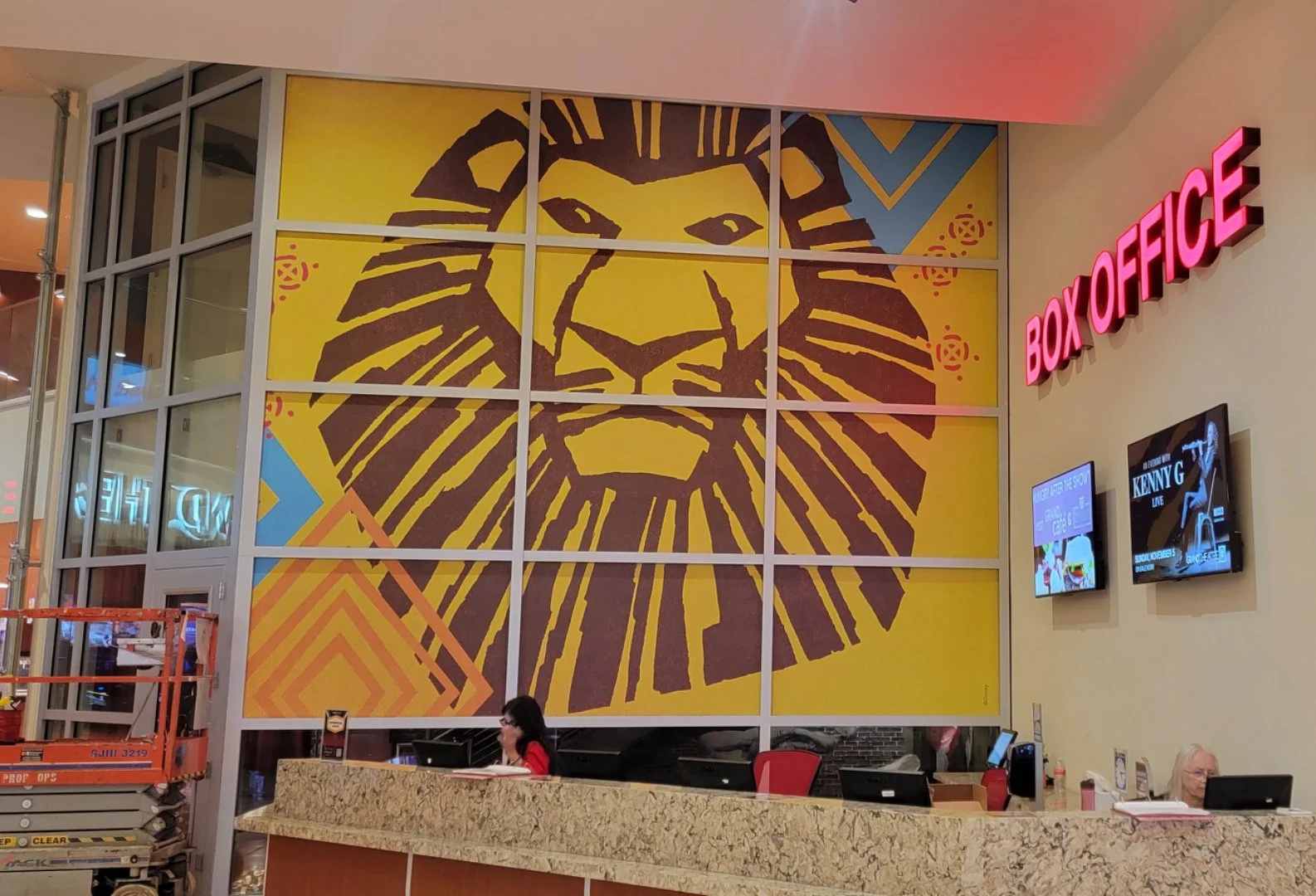 Check-in counter at a venue with a large, colorful lion's face mural on the wall behind it, and a red sign that reads 'BOX OFFICE' on the right side, with two digital screens displaying information.