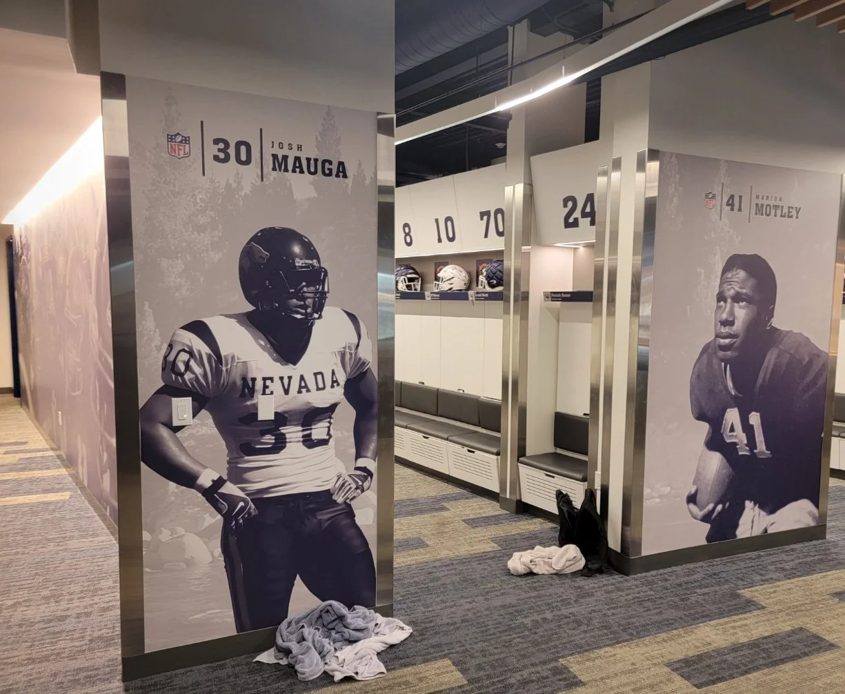 Hallway in a sports stadium with large posters of football players, including Josh Mauga from Nevada and Marlon Motley's portrait, along with lockers and helmets visible in the background.