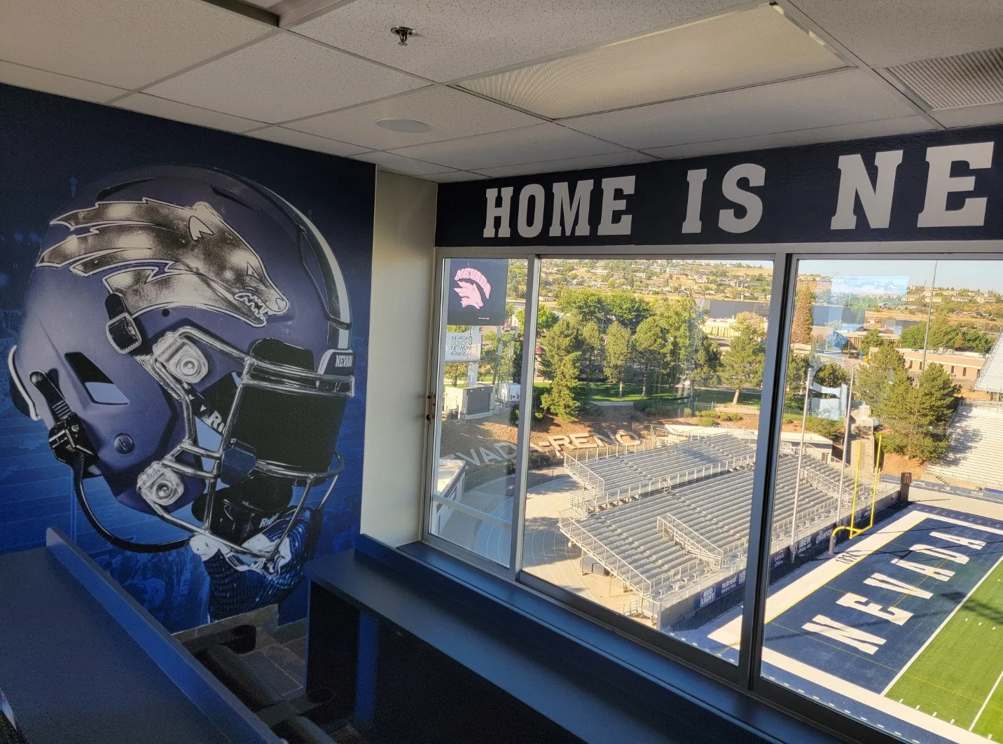 Indoor view of a sports stadium, with empty bleachers and football field, outside view through large window, mural of a sports helmet on wall, banner with text 'HOME IS NEVADA'