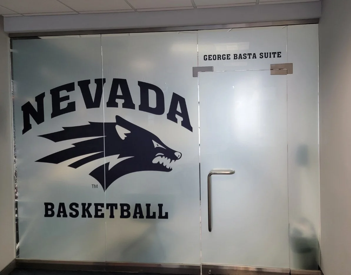 Glass door with Nevada Basketball logo and a panther mascot, labeled as 'George Basta Suite' in an indoor setting.