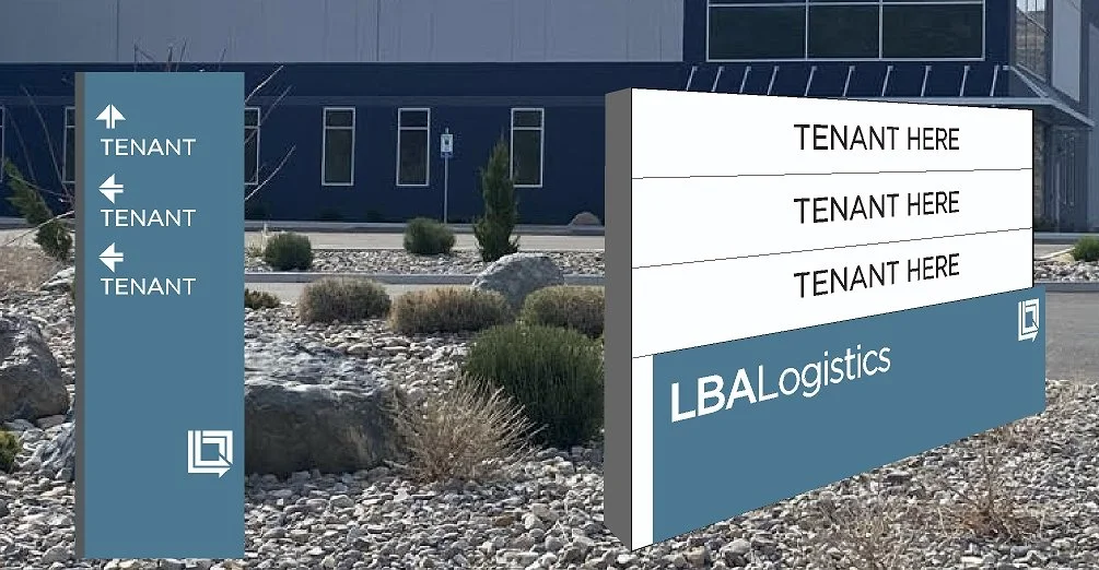A modern outdoor sign with spaces labeled for tenants and the LBALogistics company logo, situated in a landscaped area with rocks, bushes, and a building in the background.