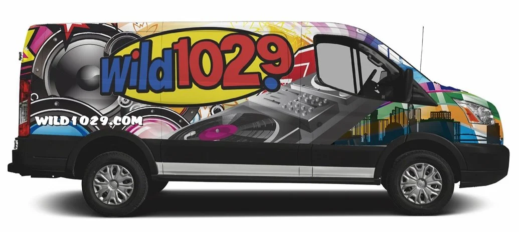 Van decorated with vibrant graphics promoting WILD102.9 radio station, featuring colorful text, speakers, DJ equipment, and city skyline imagery.