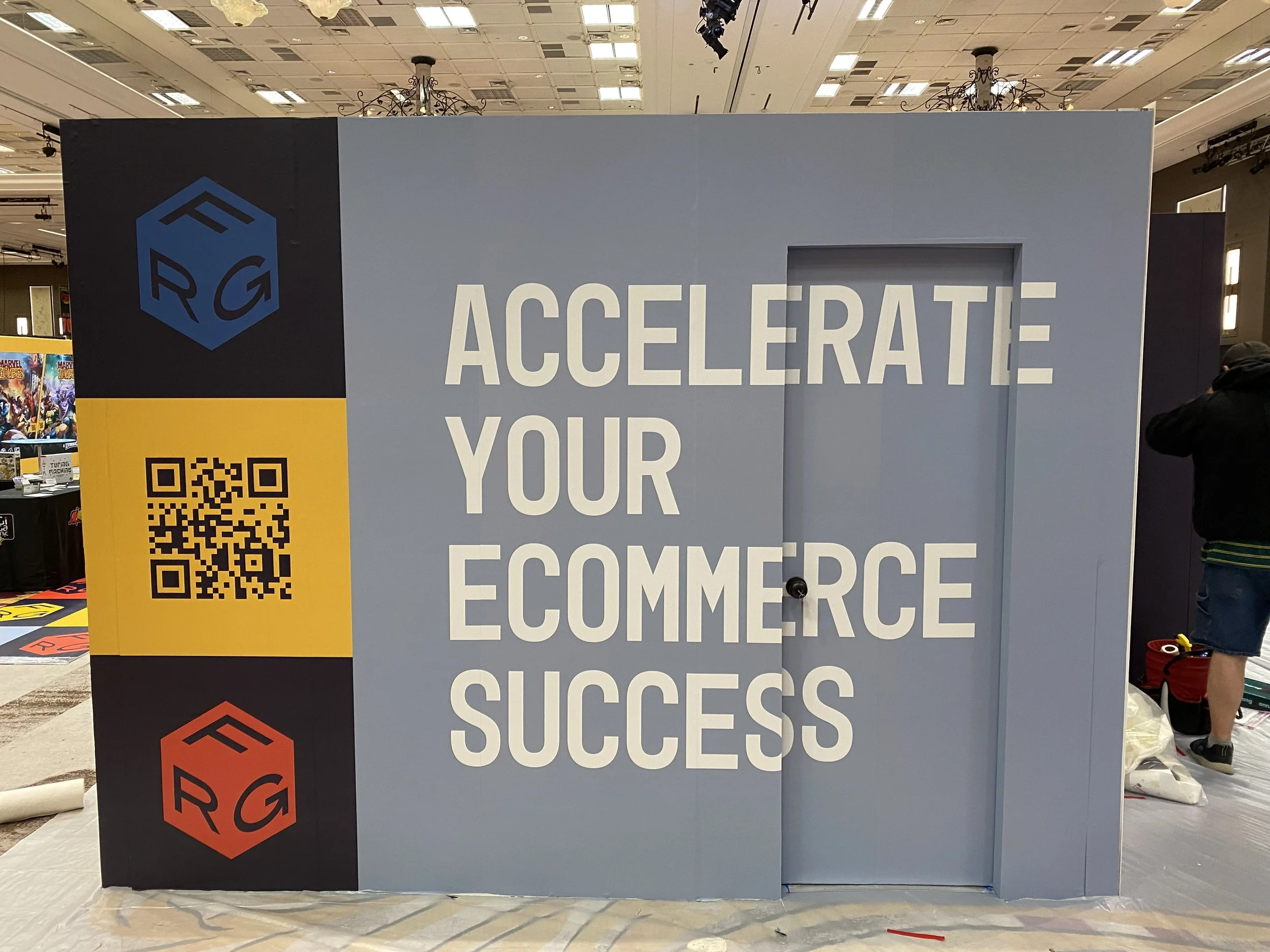 Large booth at an event with the text 'Accelerate Your E-Commerce Success'. The booth has a QR code on a yellow section on the left side, with black and red logos above and below it.