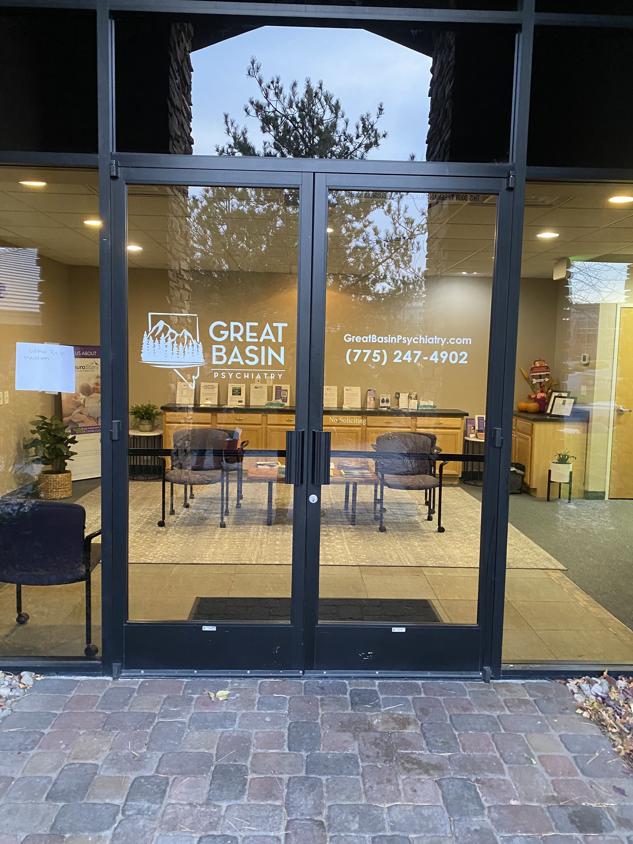 The entrance to Great Basin Psychiatry with glass double doors displaying the name, website, and phone number. Inside, there are chairs, a reception desk, and potted plants.