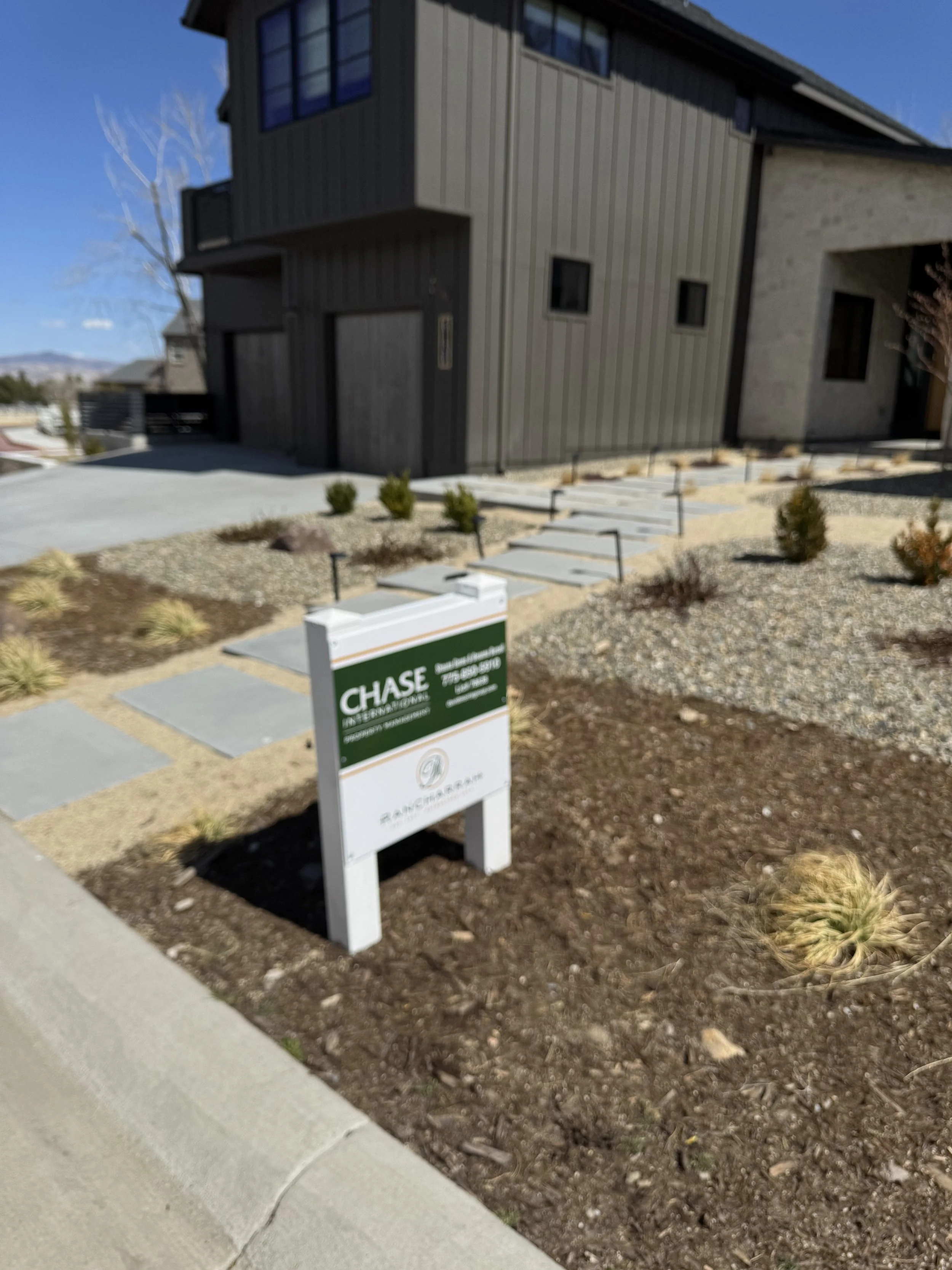 A for sale sign in front of a modern house with a landscaped yard featuring small bushes, gravel, and stepping stones.