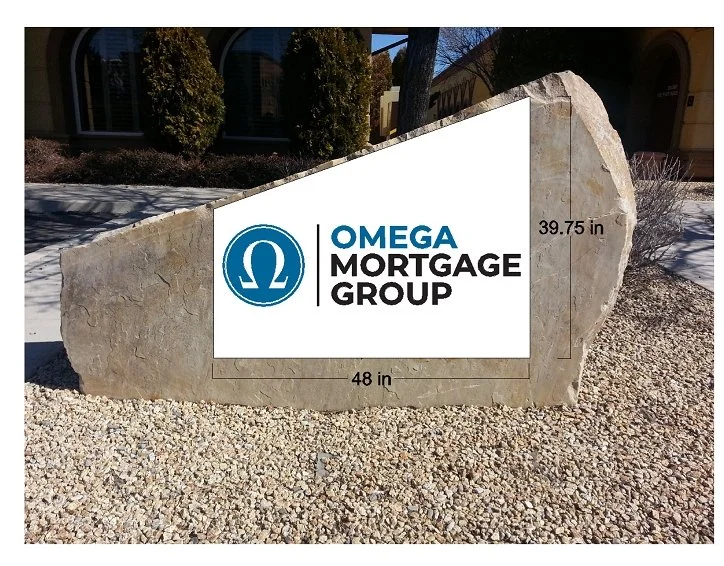 A large stone sign with the logo and text for Omega Mortgage Group, located outdoors on a bed of gravel with trees and buildings in the background.