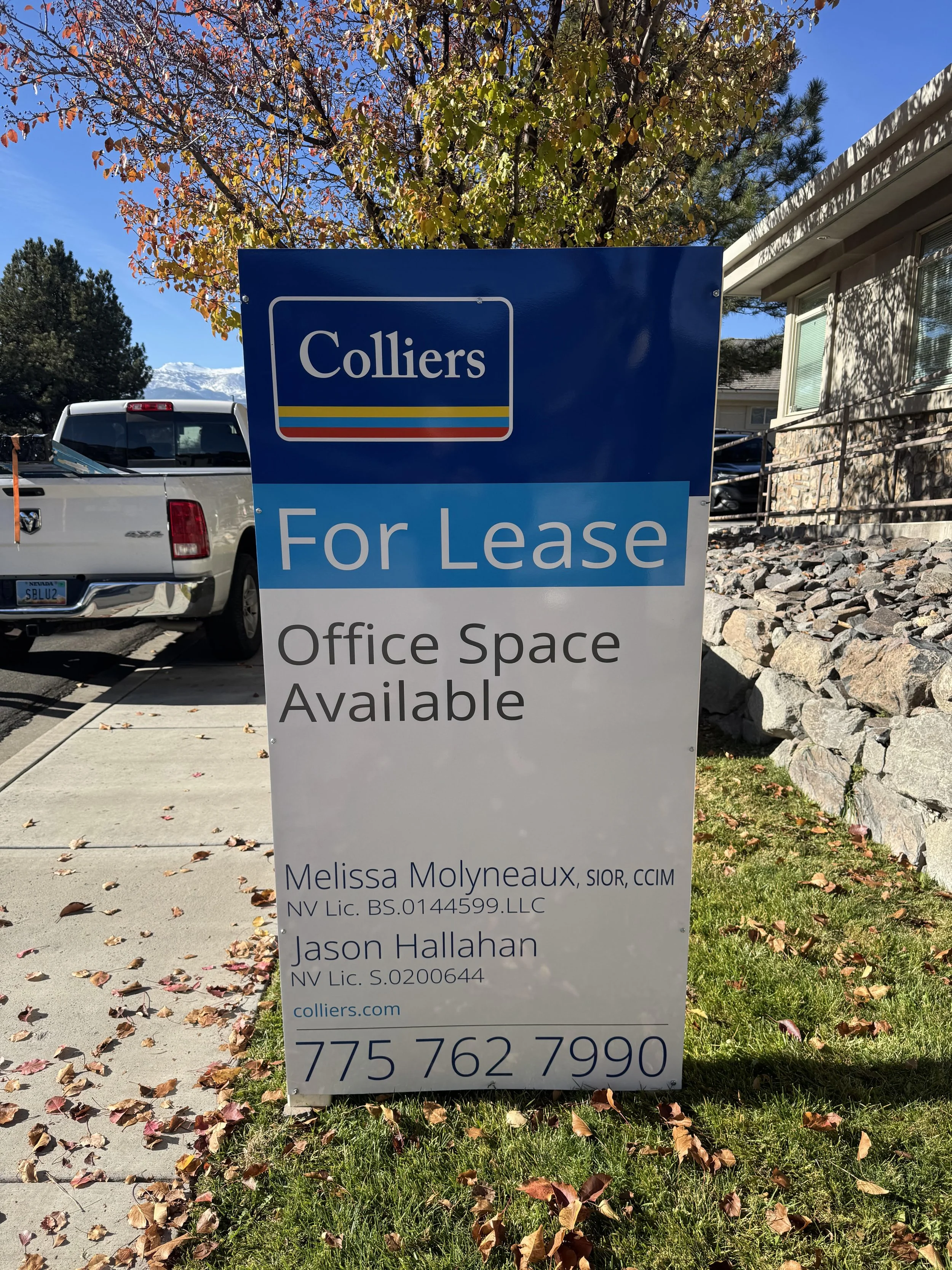 Sign advertising office space for lease by Colliers, with contact information and agents' names, set outdoors on a sidewalk with fallen leaves, trees with autumn foliage, and a house with large windows and stone landscaping in the background.