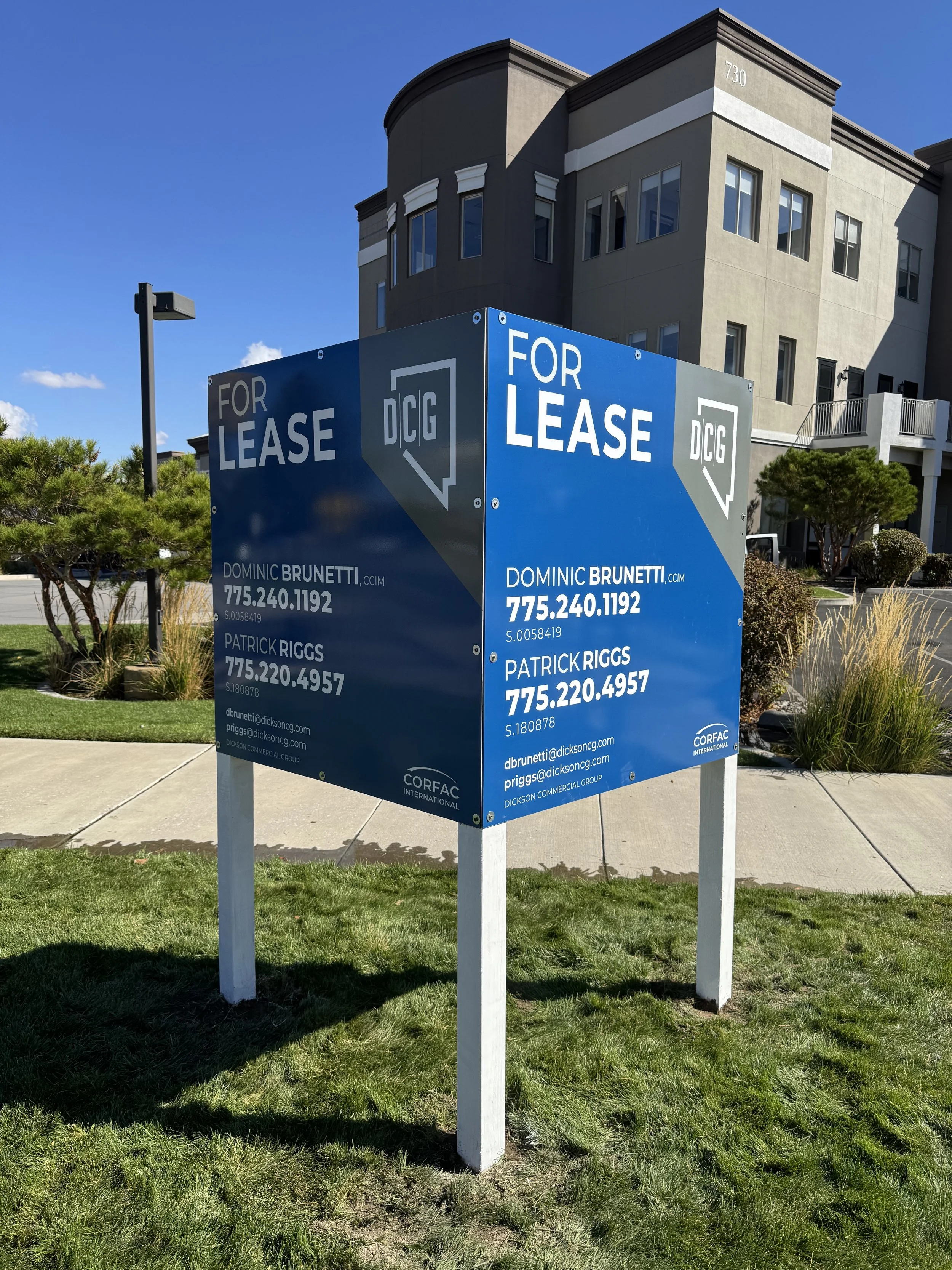 A blue sign with white text reading "For Lease" and contact information for Dominic Brunetti and Patrick Riggs. The sign is in front of a modern multi-story residential building with grey and beige exterior, large windows, and landscaped surroundings