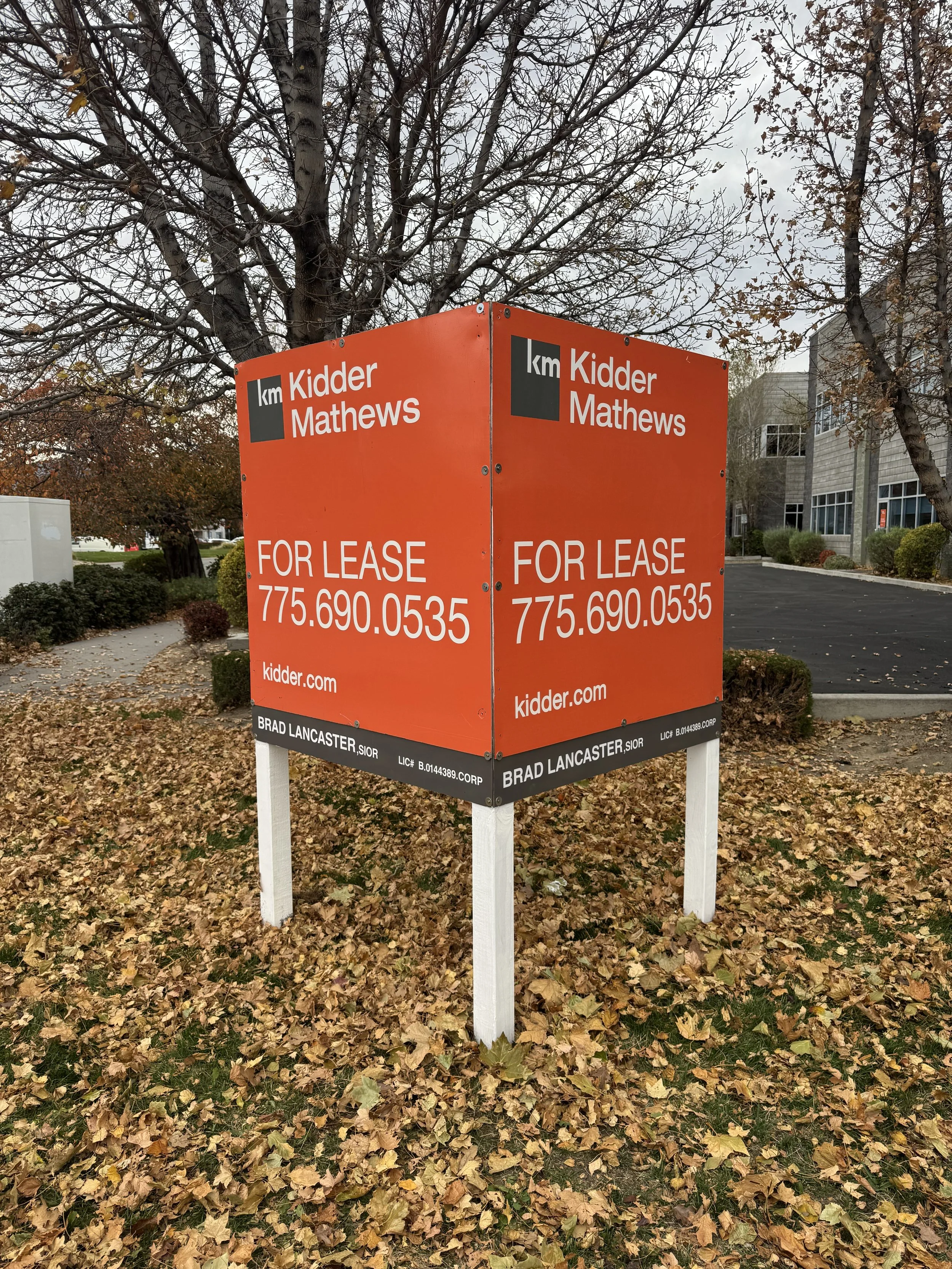 An outdoor advertisement display with a bright orange background that promotes a property for lease, displaying a contact phone number and website, positioned on white wooden posts in a park-like area with fallen leaves, leafless trees, and nearby bu