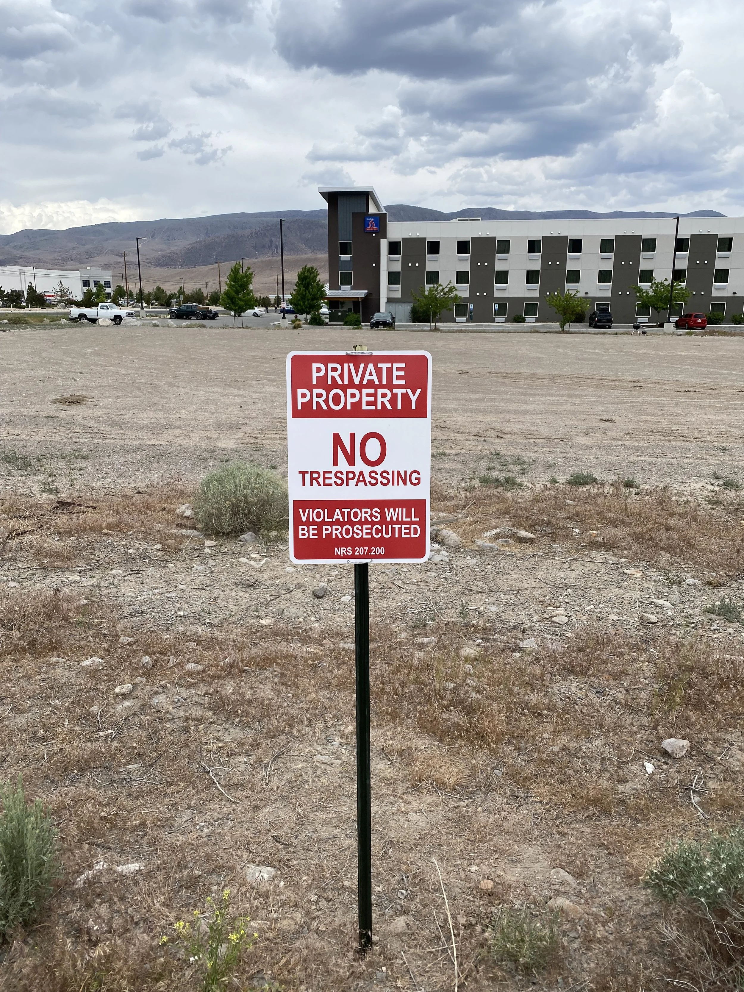 A sign on a patch of dry, rocky ground states 'Private Property, No Trespassing, Violators Will Be Prosecuted,' with a background of a large, modern building and a parking lot under a cloudy sky.