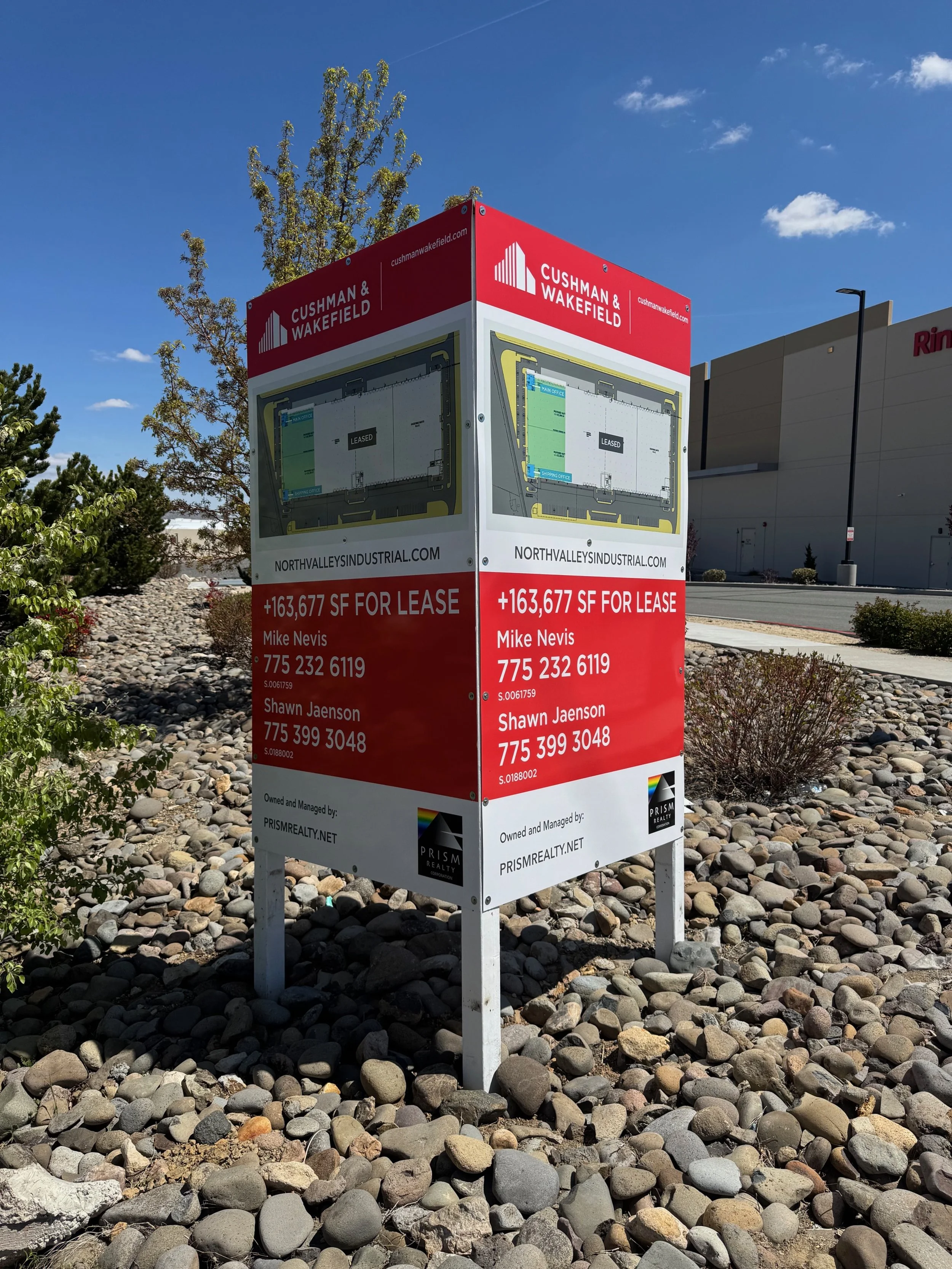 Outdoor sign advertising available industrial space for lease, featuring an illustrated floor plan, contact information, and company logos, surrounded by rocks and bushes with a building, trees, and blue sky in the background.