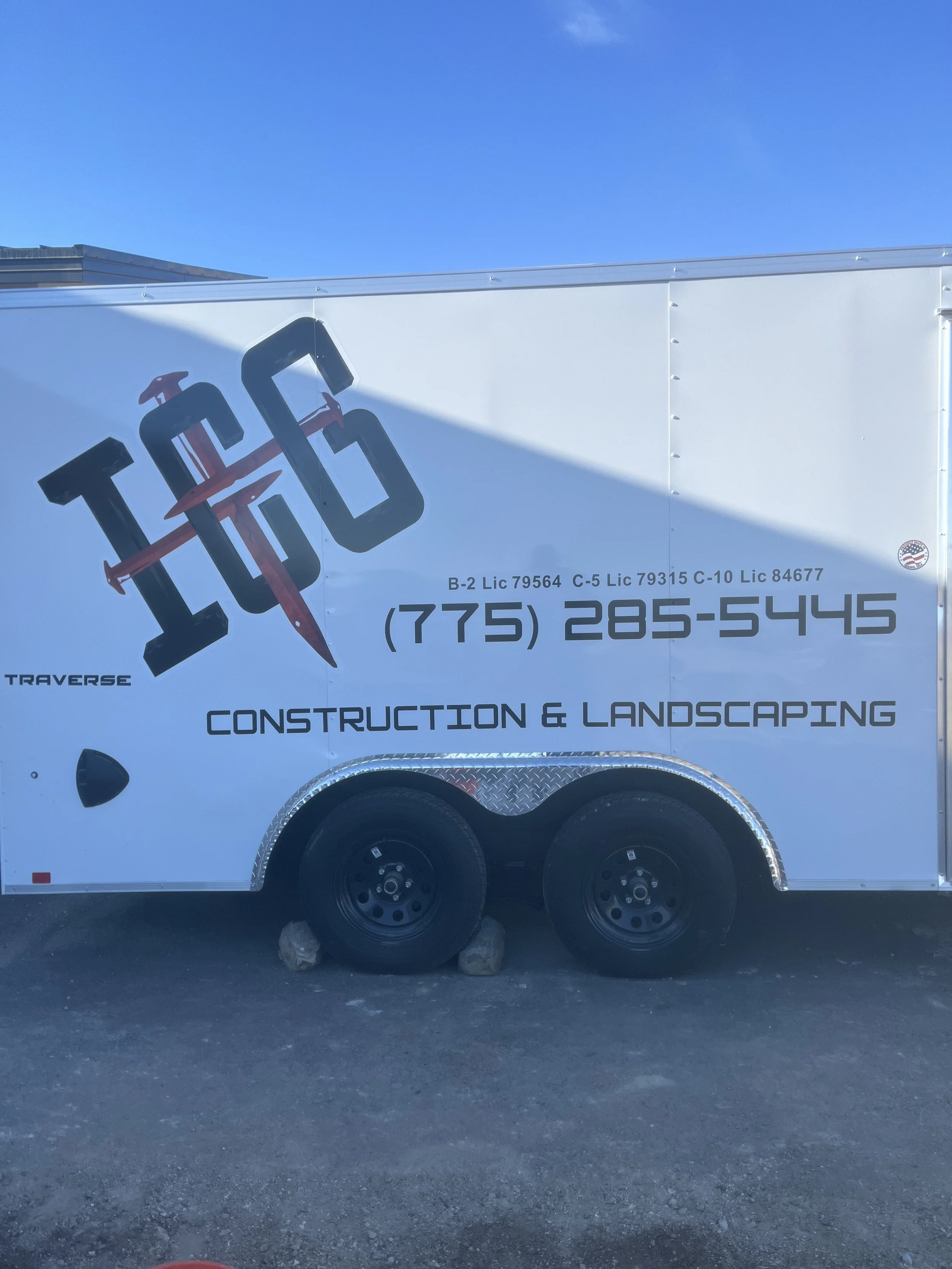 A white construction trailer with black and red logo and text showing a phone number, license numbers, and the words "Construction & Landscaping".