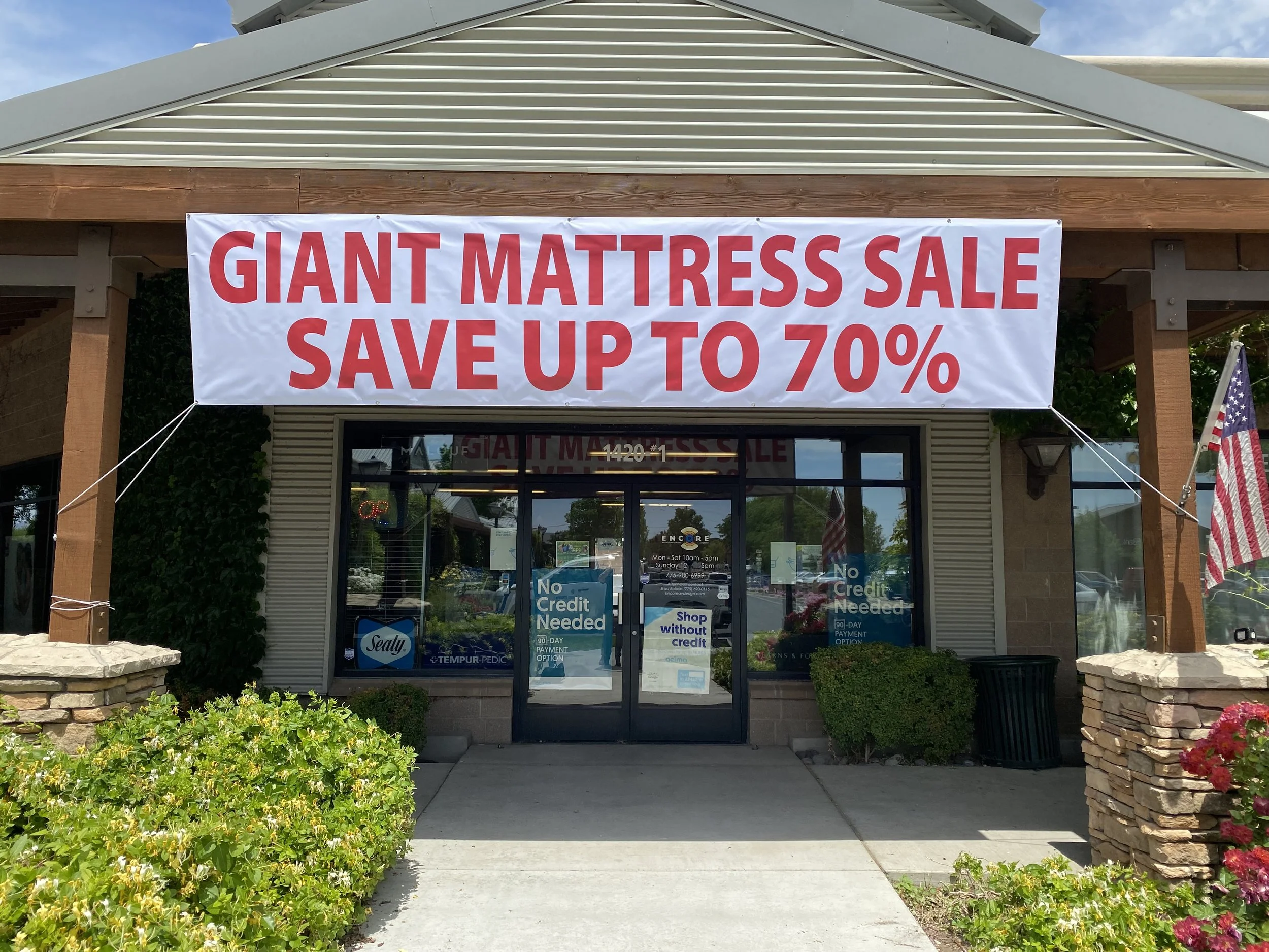 Large banner reading 'Giant Mattress Sale Save Up to 70%' hanging above the entrance of a store with glass doors and windows, surrounded by greenery and stone planters.