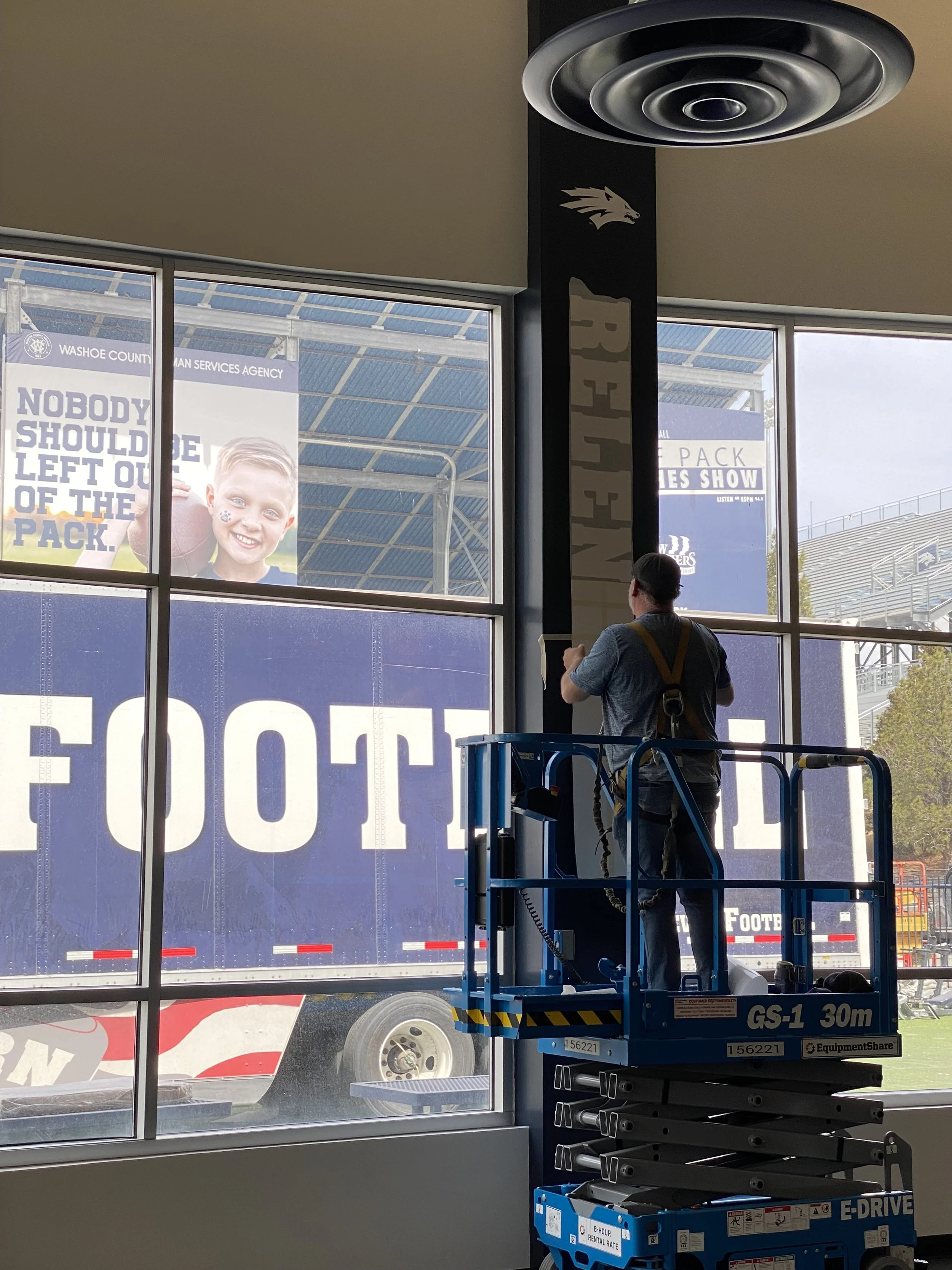 A worker on a blue lift inside a building, cleaning or installing a large window with a view of a football stadium outside.