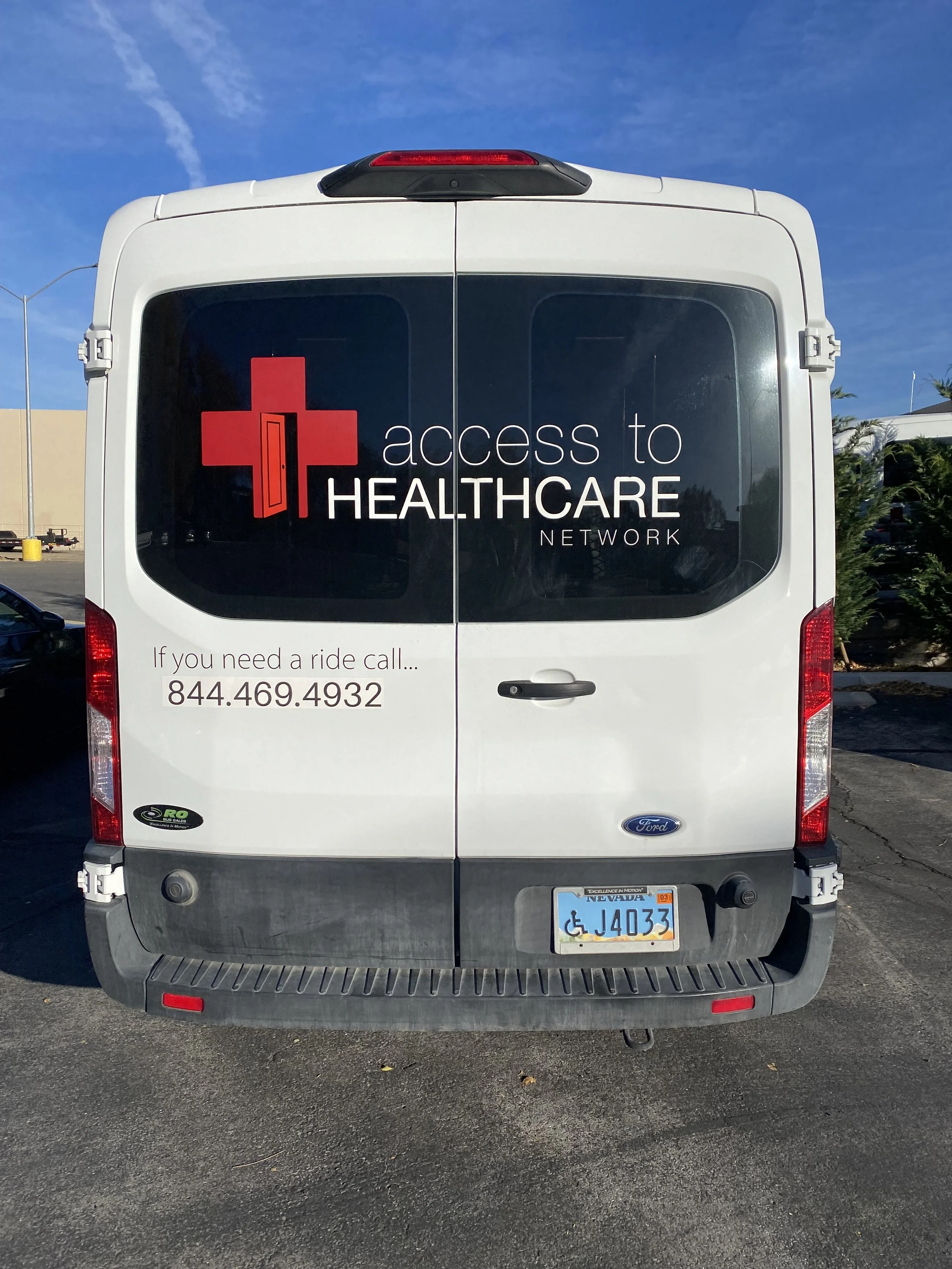 The back of a white Ford van with a logo for Access to Healthcare Network, a red cross with a door silhouette, and text providing a contact number for rides services.