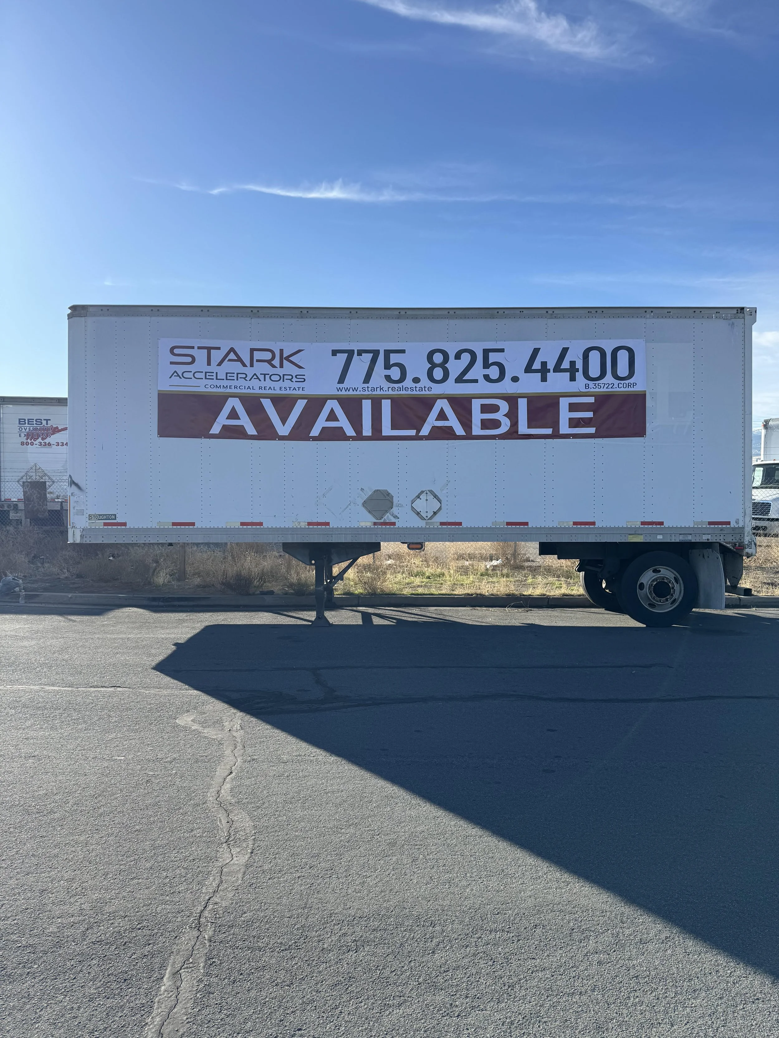 A large white trailer with a sign that reads 'Stark Accelerators, Commercial Real Estate' and a phone number, with a banner below that says 'Available.' The trailer is parked on a paved lot with a partially sunny sky overhead, and other trucks are vi