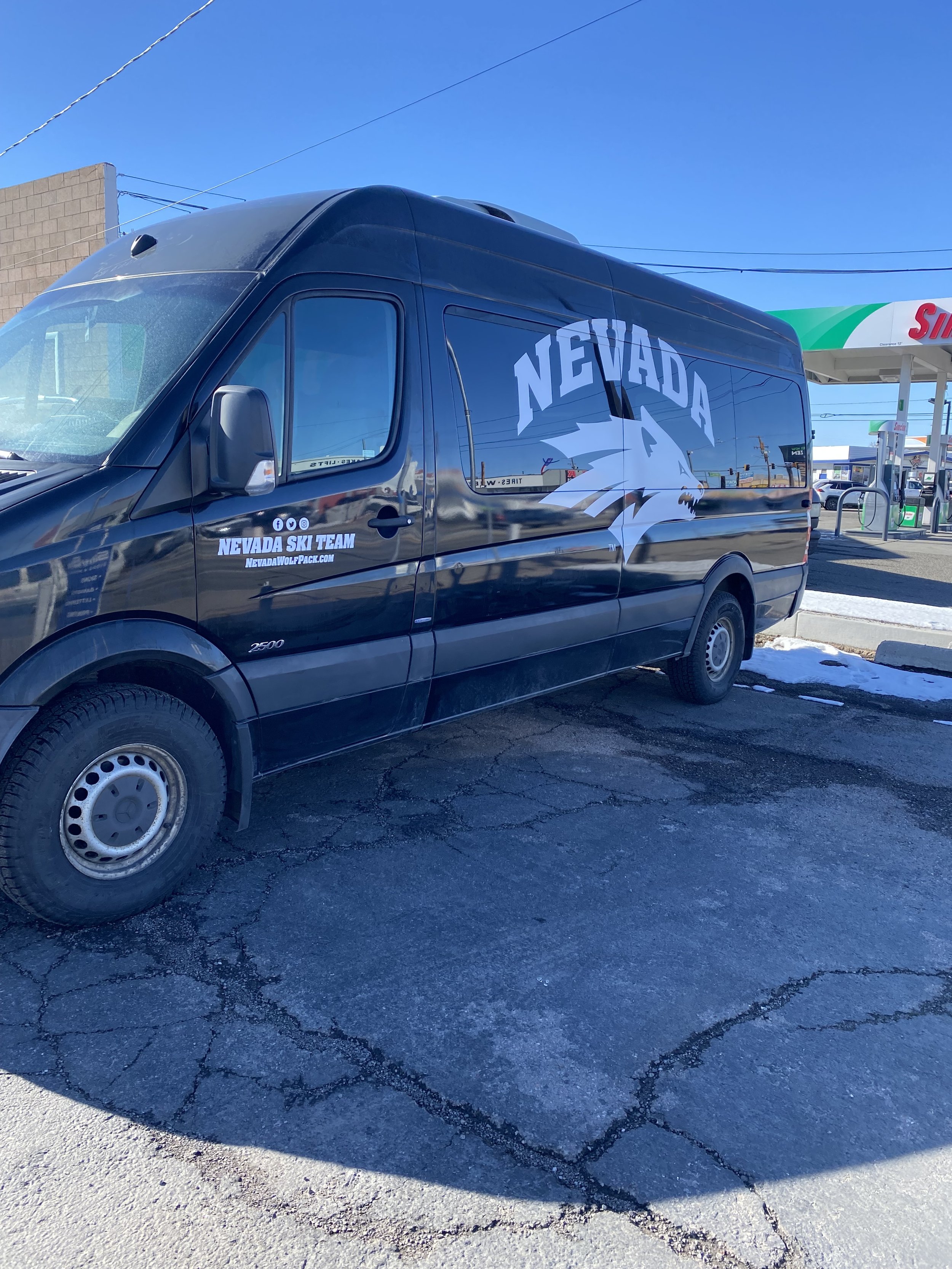 Black van with Nevada ski team logo and wolf graphic parked at a gas station with a Shell sign, with snow on the ground and a clear blue sky.
