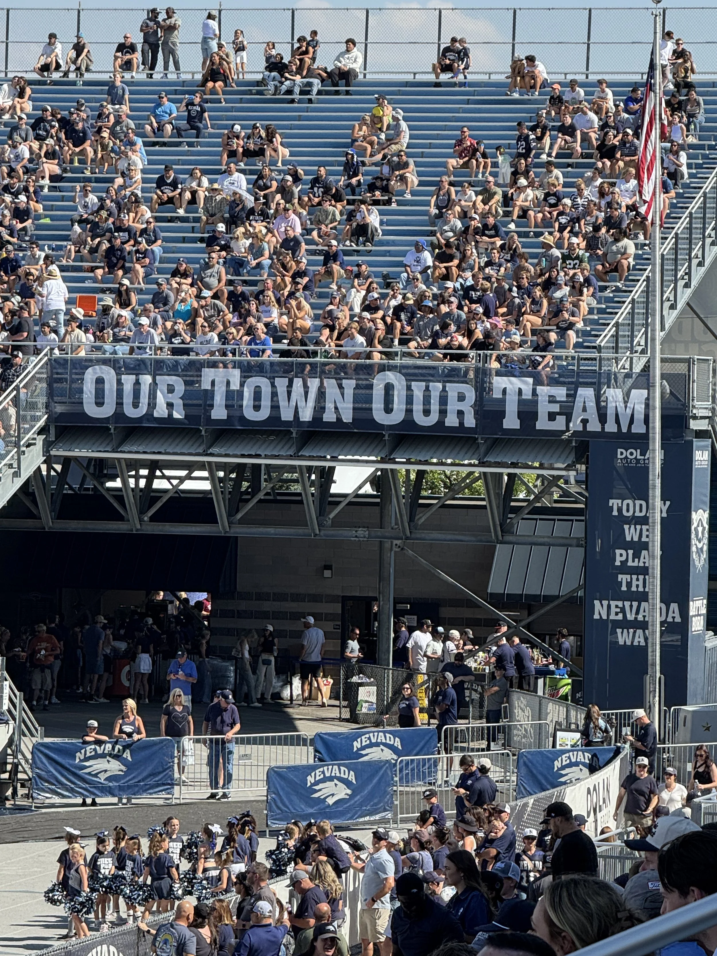Crowd of people seated in bleachers and walking around at an outdoor event, with a banner that reads 'Our Town Our Team' and Nevada state flags.