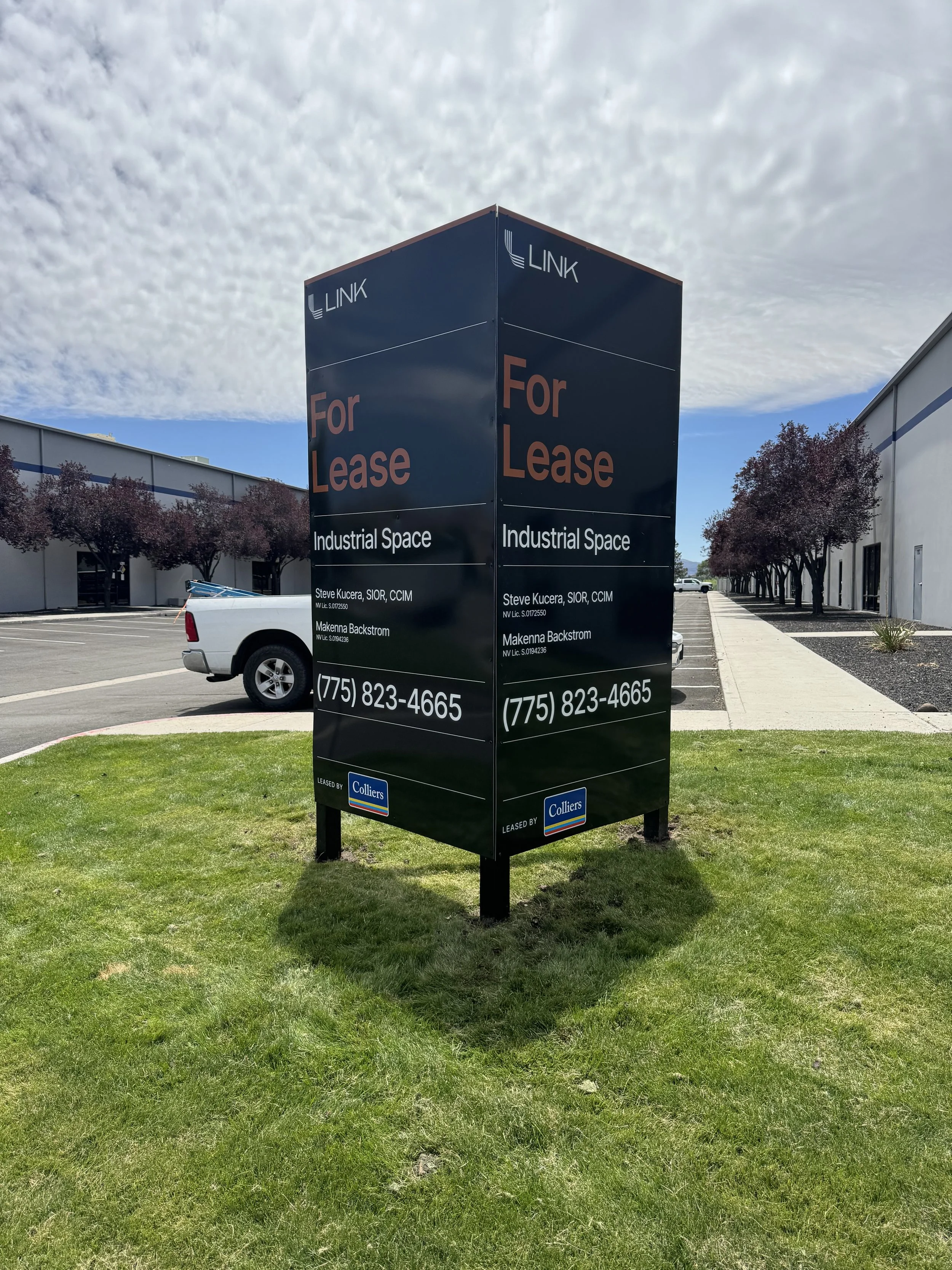 A large black sign in a grassy area advertising industrial space for lease, with contact information and a leasing company logo, beside a parking lot and commercial buildings.