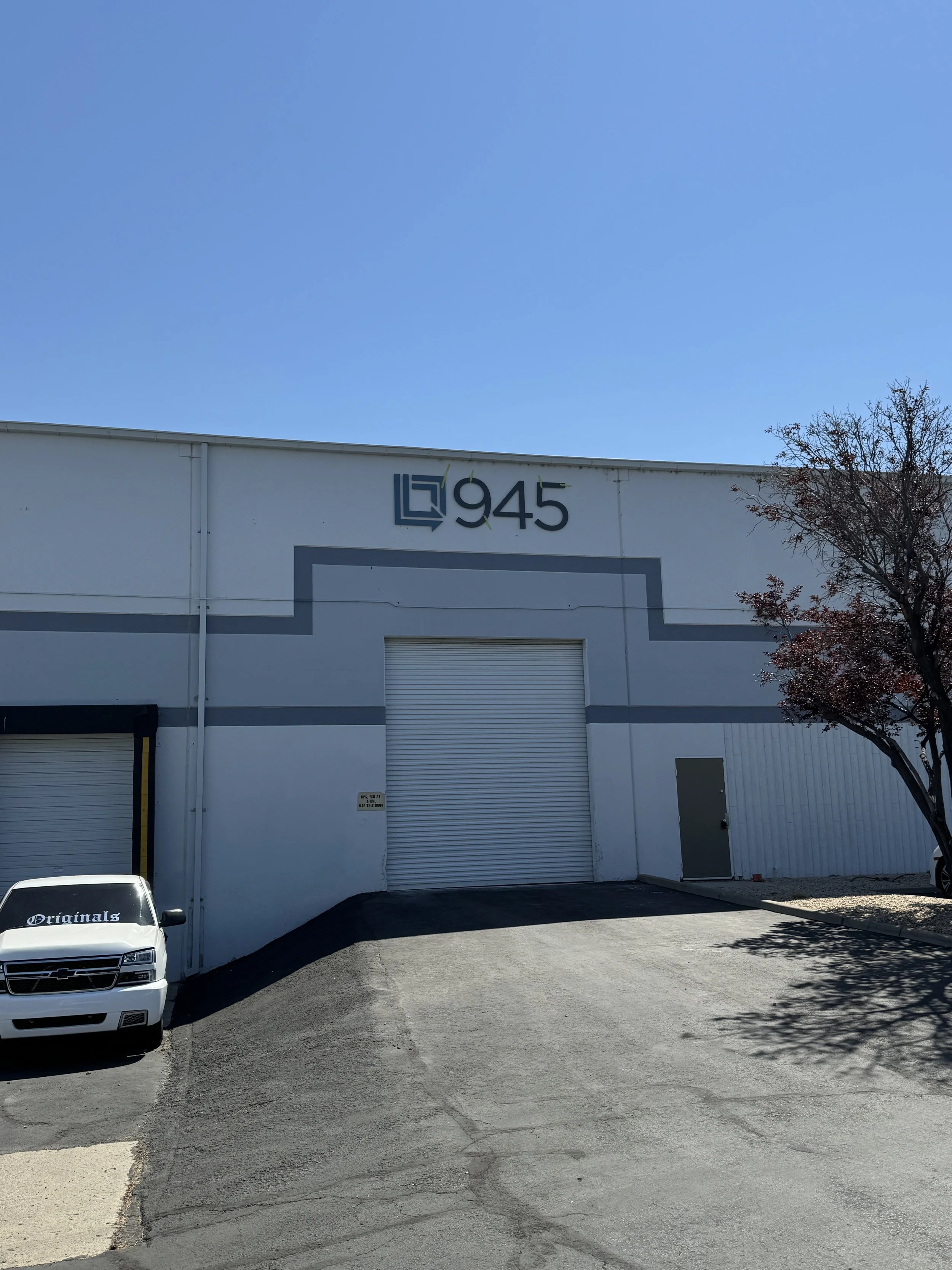 Industrial building with the number 945 and a logo above it, a driveway in front, a white truck parked on the left, a tree on the right, and a clear blue sky.