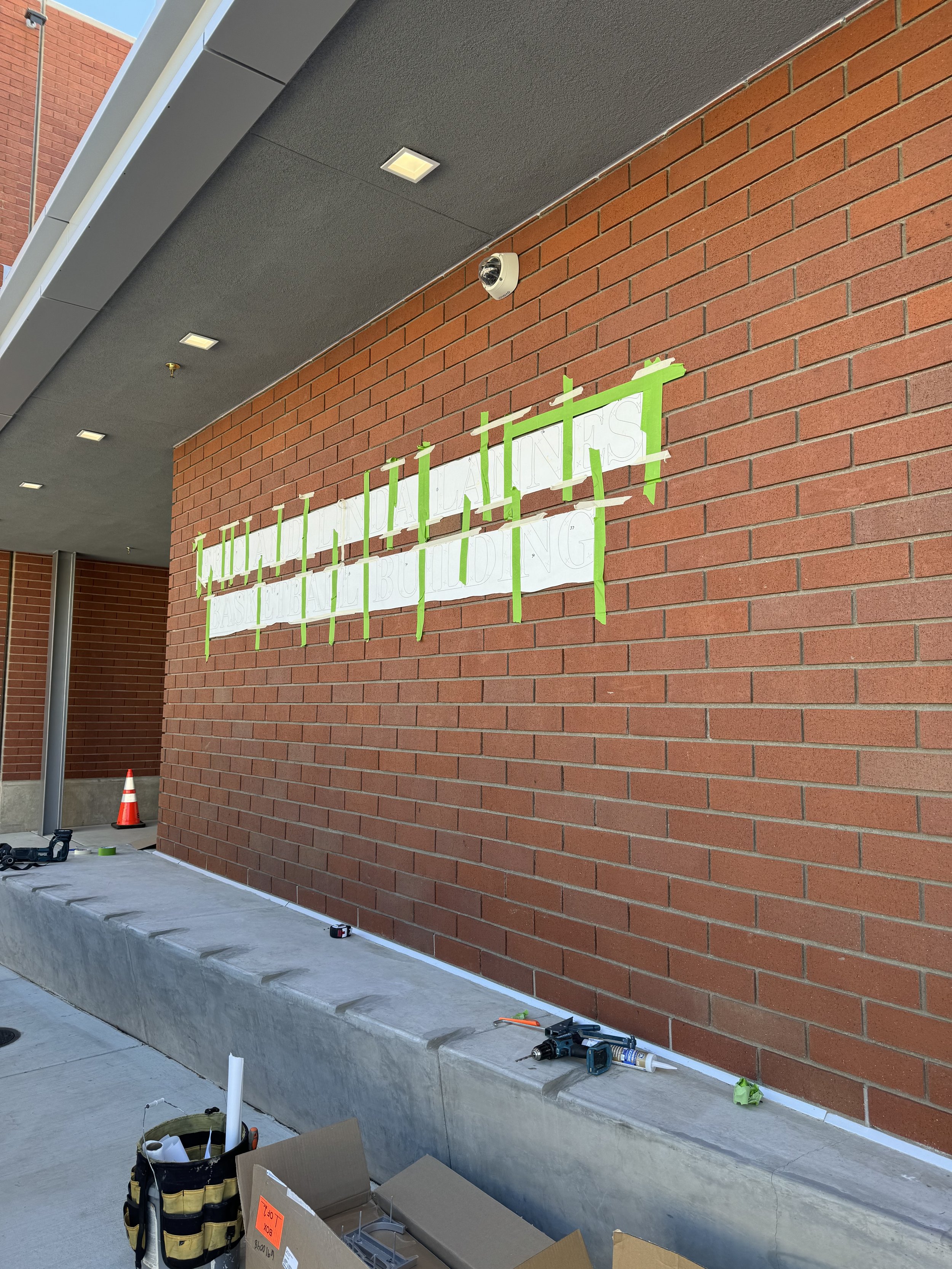 A brick building wall with a partially revealed sign that reads 'NAILAITAS.' The sign is taped on with green painter's tape. Construction tools and supplies are on the sidewalk below, and there is a cone on the left side.