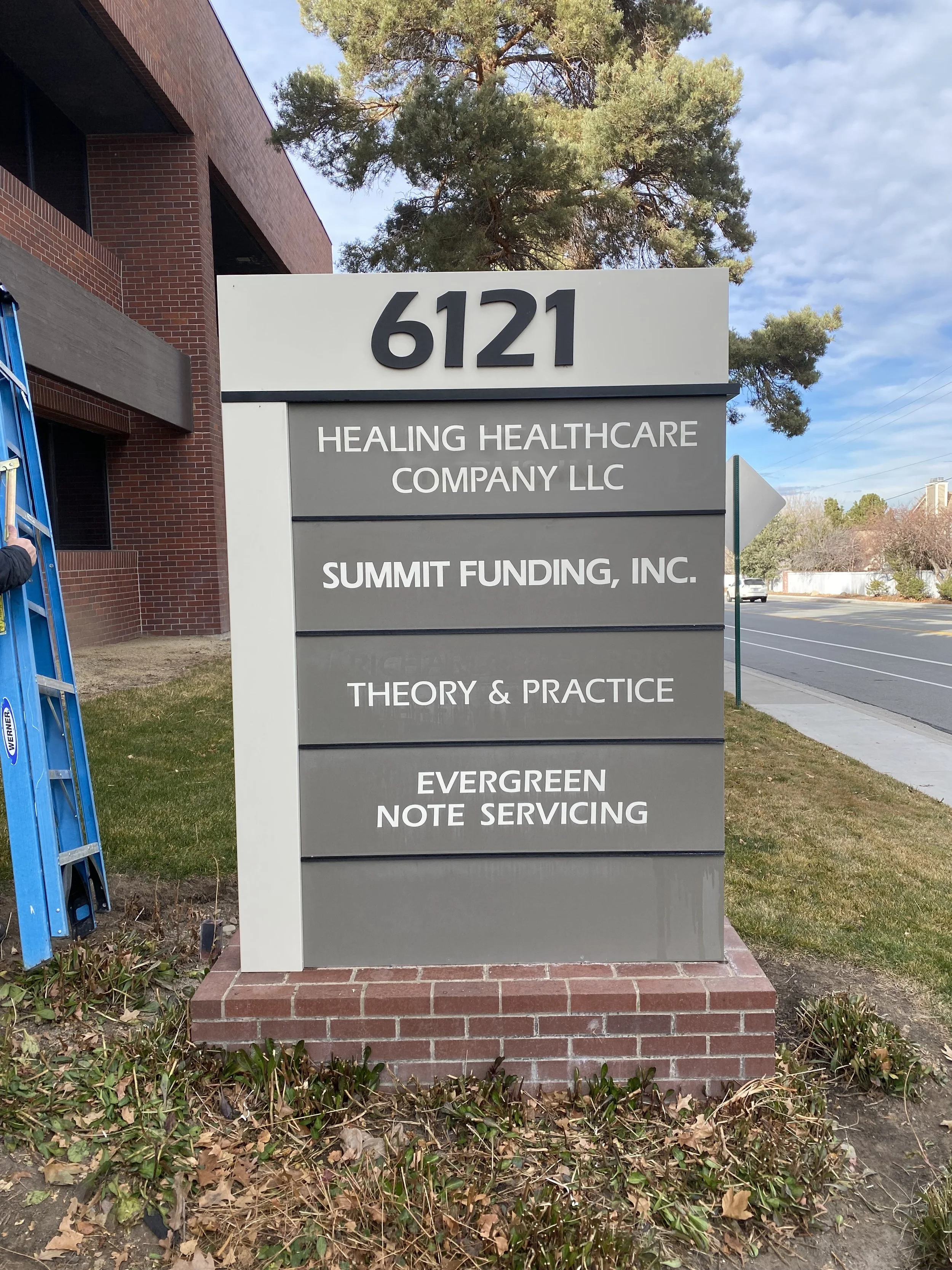 Sign with the address 6121 and listing four businesses: Healing Healthcare Company LLC, Summit Funding Inc., Theory & Practice, Evergreen Note Servicing, on a grassy area near a sidewalk with trees and a street in the background.
