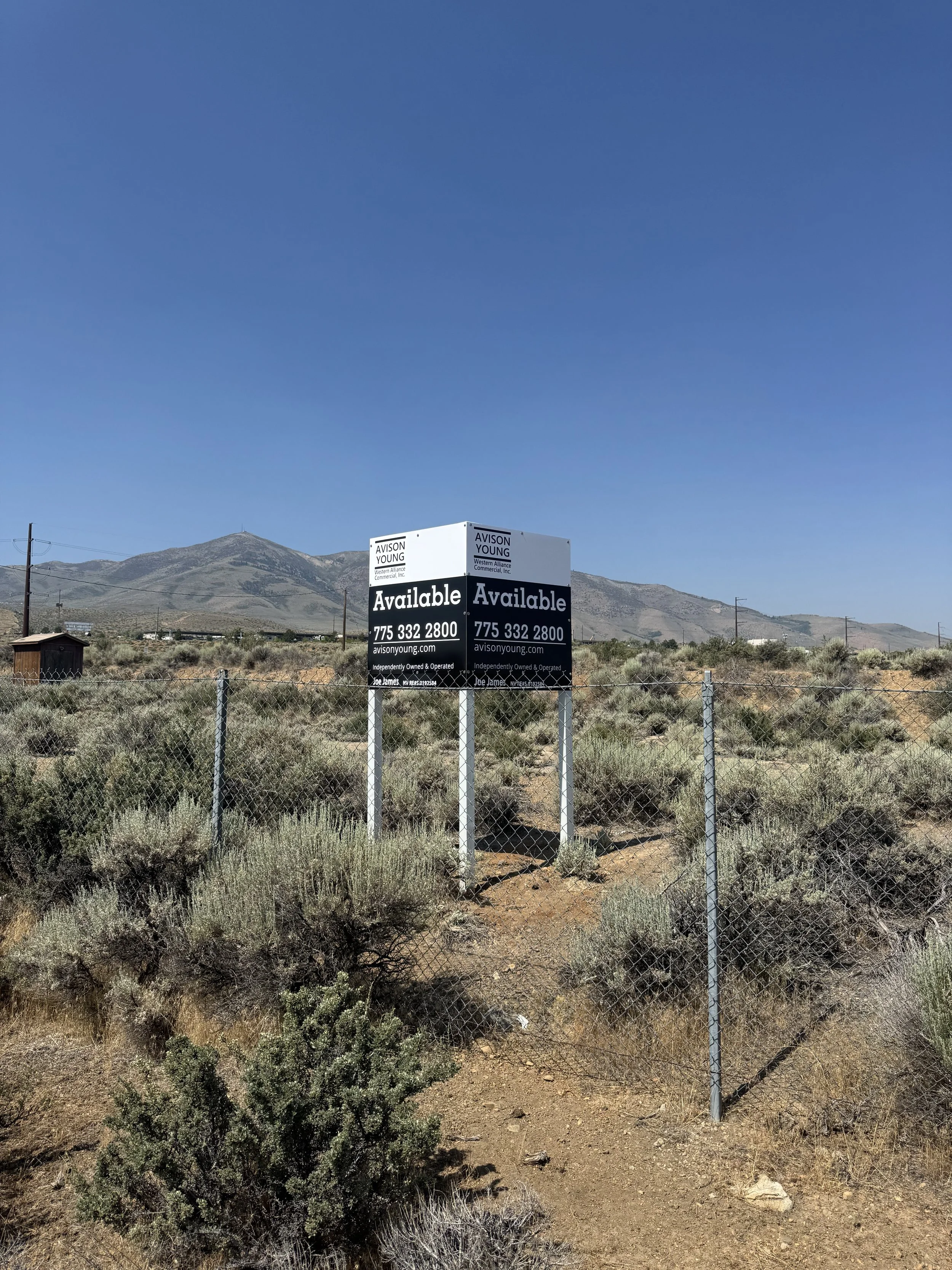 A chain-link fence with an 'Available' real estate sign in a dry, desert landscape with hills or mountains in the background.