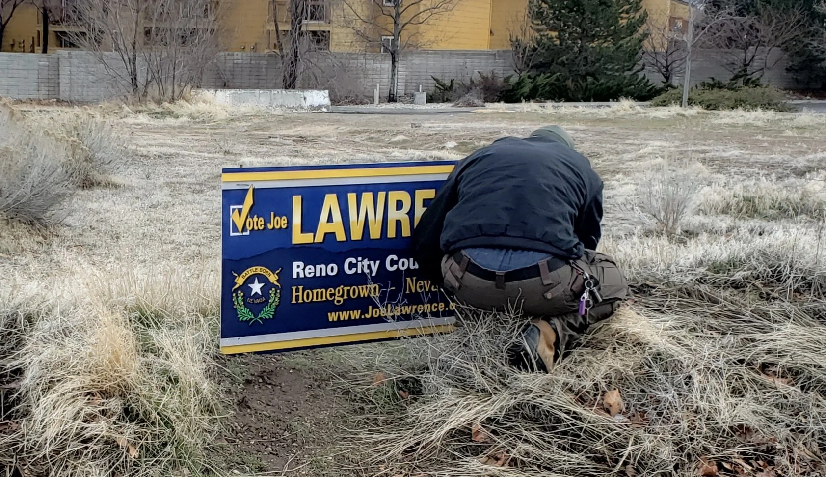 A sign worker kneeling on dry grass beside a political campaign sign that reads 'Vote Joe LAWRENCE' for Reno City Council. The sign includes the text 'Homegrown Nevada' and a website URL.