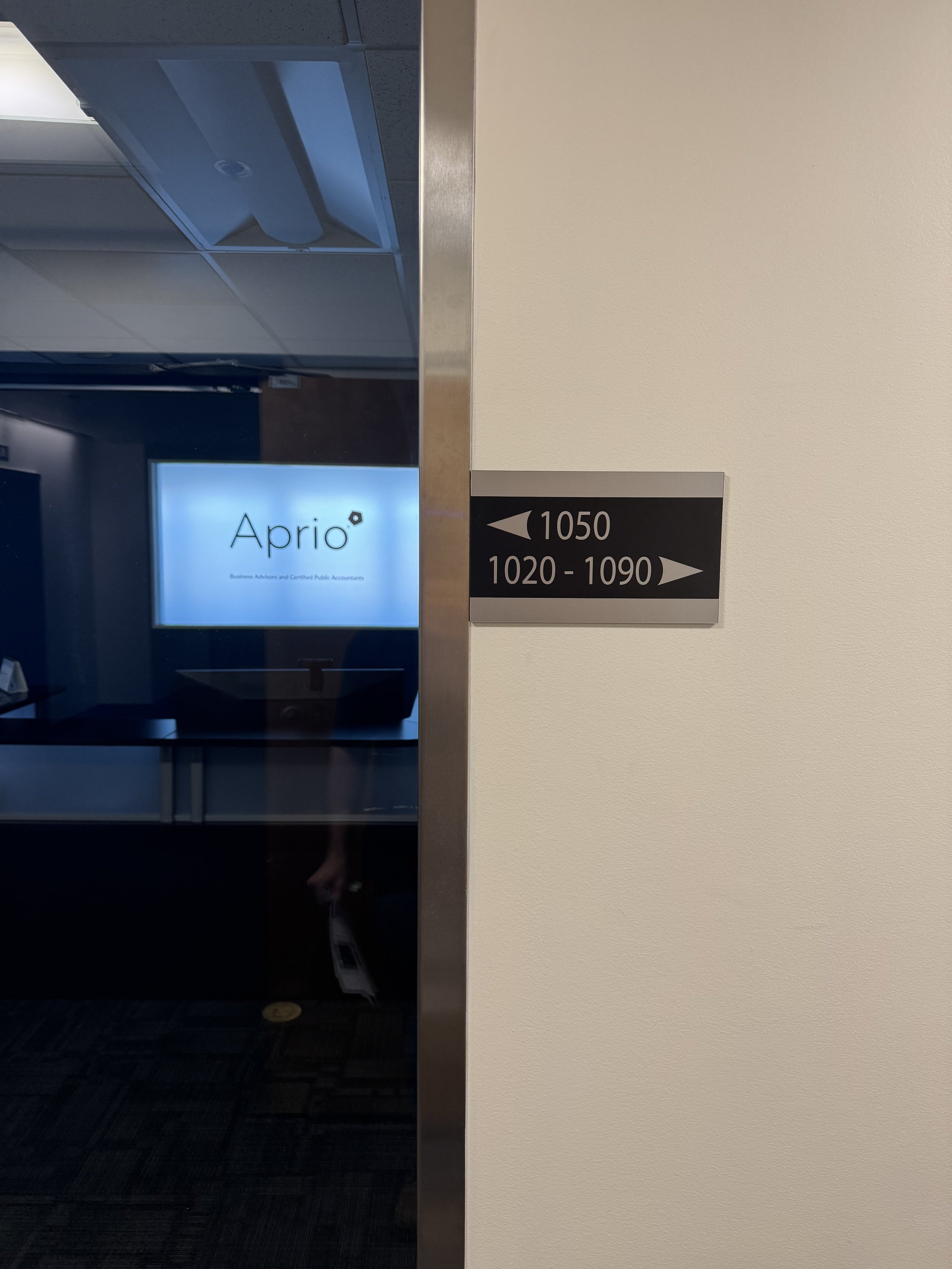 Office directory sign with room numbers 1050 to the left and 1020 to 1090 to the right, and a glass wall showing the Aprio logo and office interior.