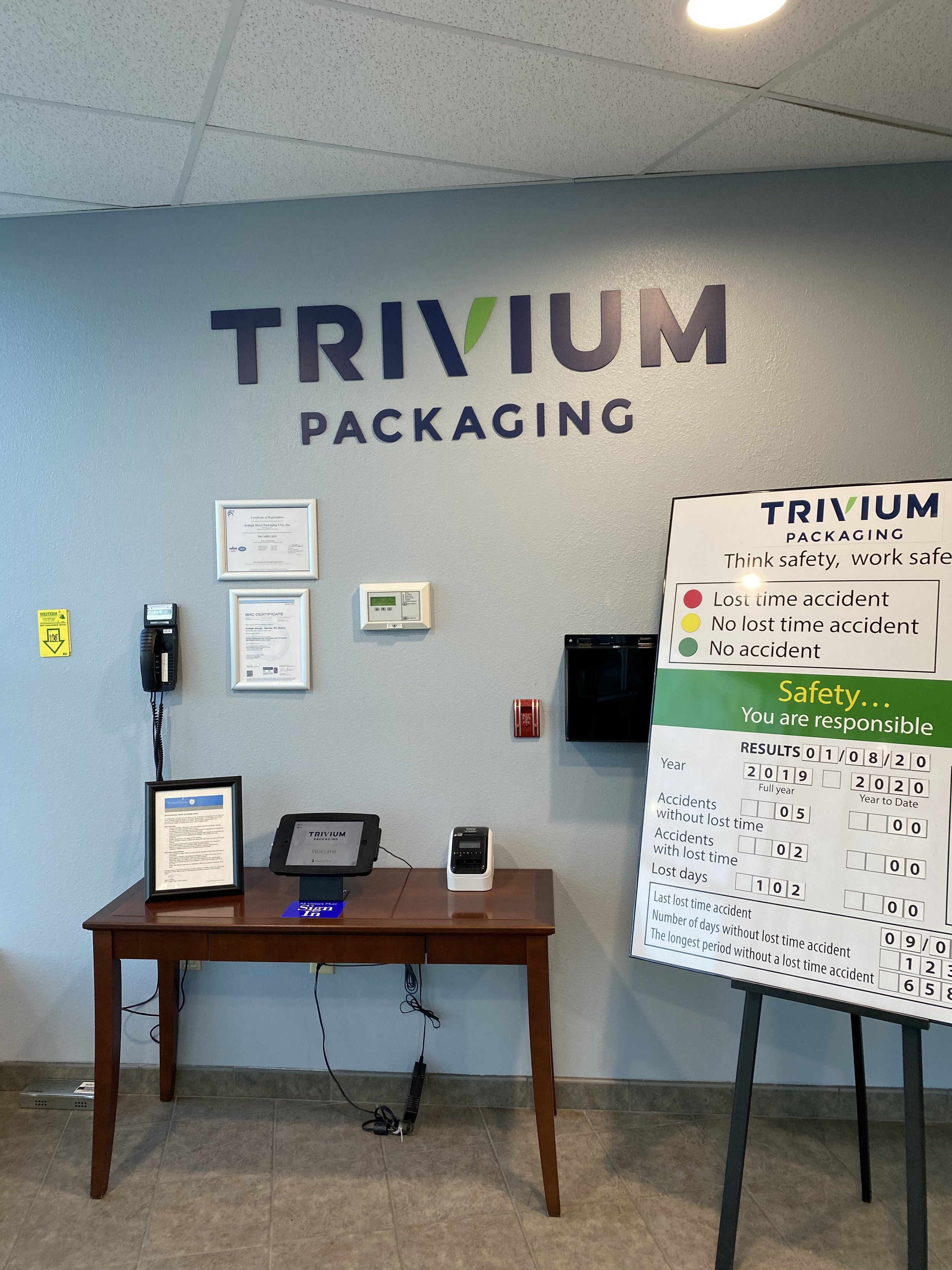 Office wall with 'TRIVIUM PACKAGING' logo, framed certificates, a wall-mounted phone, a thermostat, and a safety and accident report board on an easel. A small wooden desk underneath the wall has a framed document, a tablet device, and a phone.