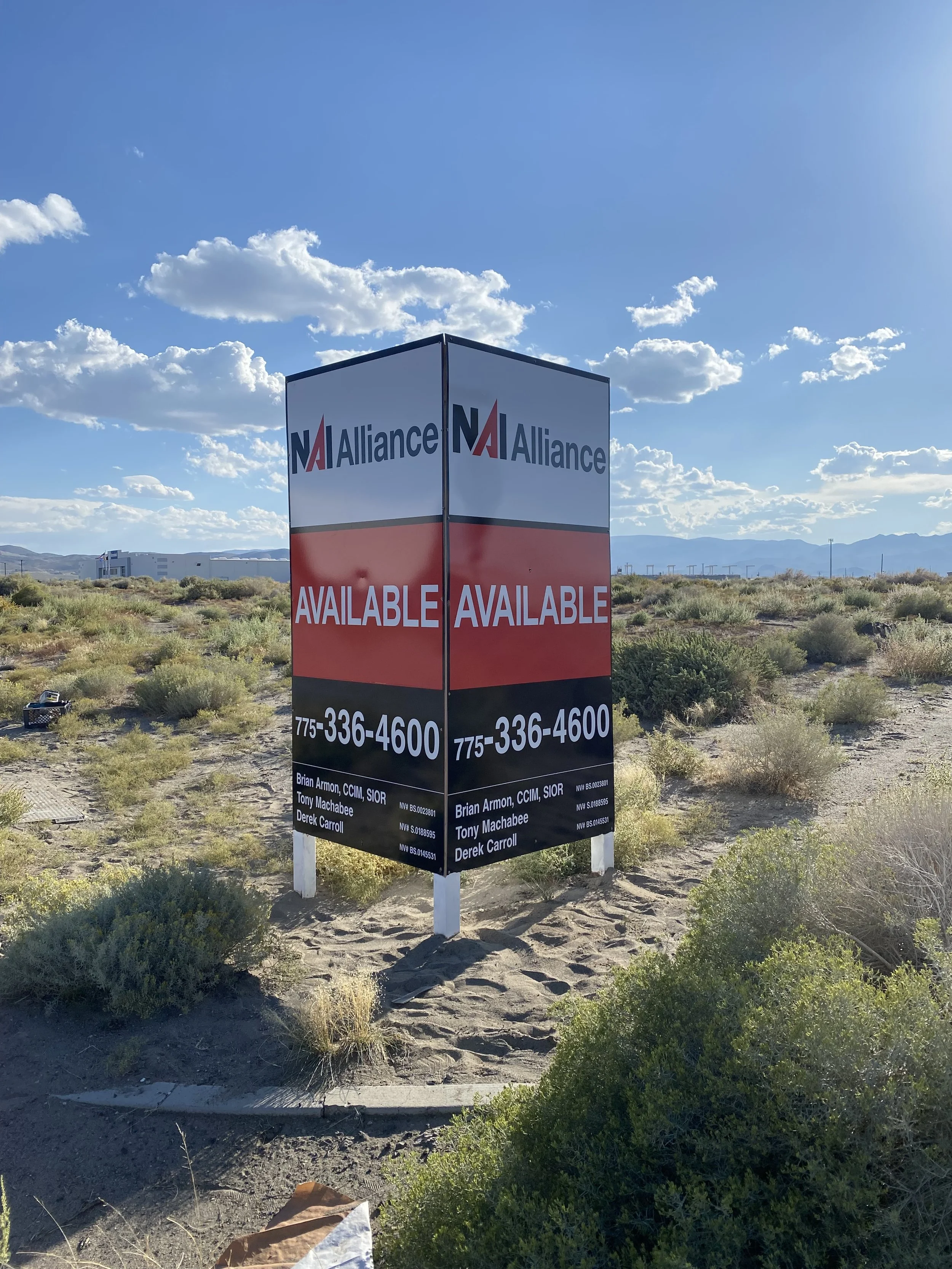 A roadside sign in a desert landscape advertising availability for real estate, with the words 'NA Alliance,' 'Available,' and a phone number.