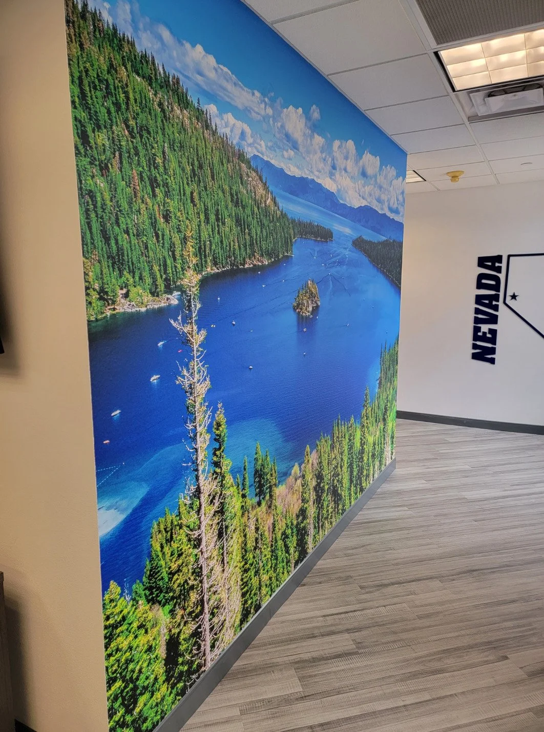 Large wall mural depicting a scenic view of a lake surrounded by dense evergreen forest, with mountains in the background under a partly cloudy sky. The scene includes several boats on the water and a small island in the lake.