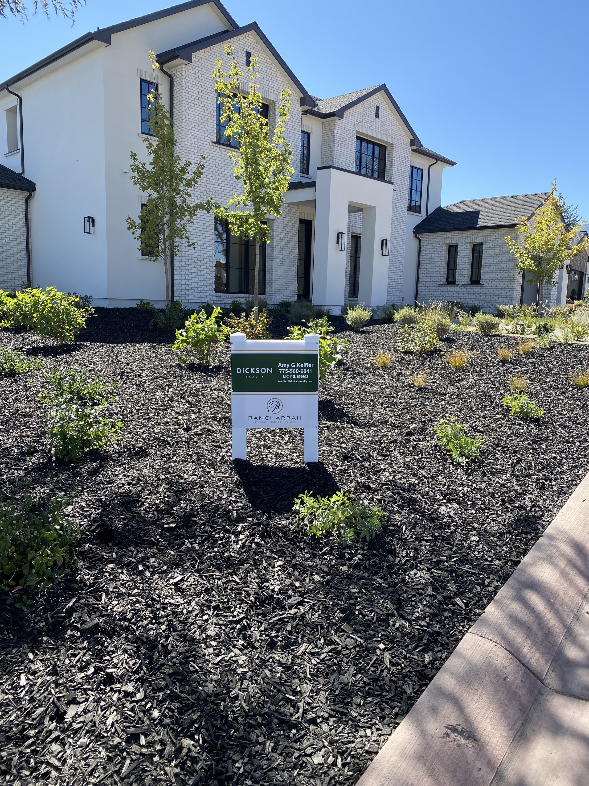 Newly built white brick multi-story house with black window frames, surrounded by young trees and shrubs, and a front yard with dark mulch. A sign in the yard displays real estate agent contact information.