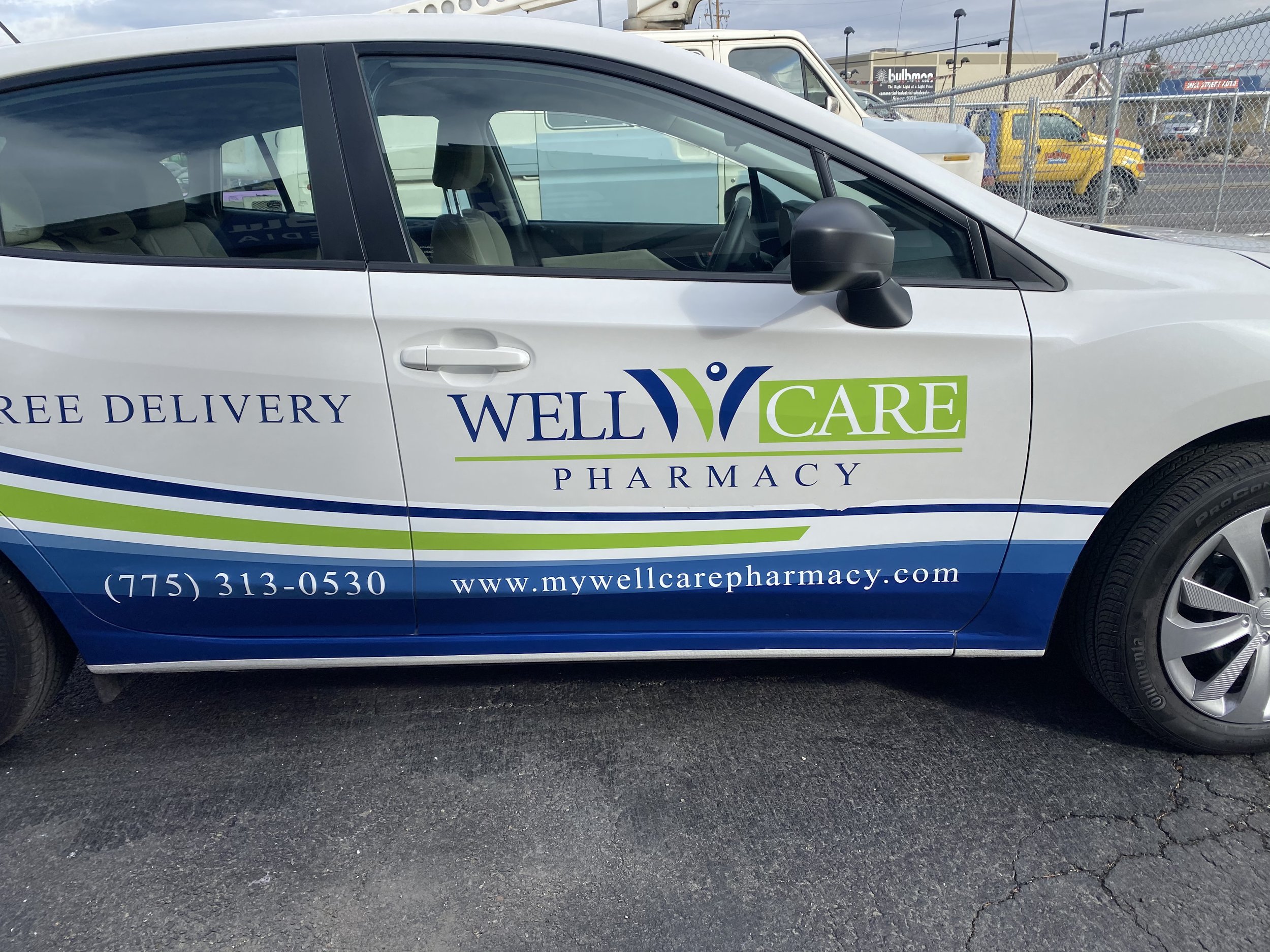 Side view of a white and blue Well Care Pharmacy vehicle with contact information and website, parked on asphalt with a chain-link fence and other vehicles in the background.