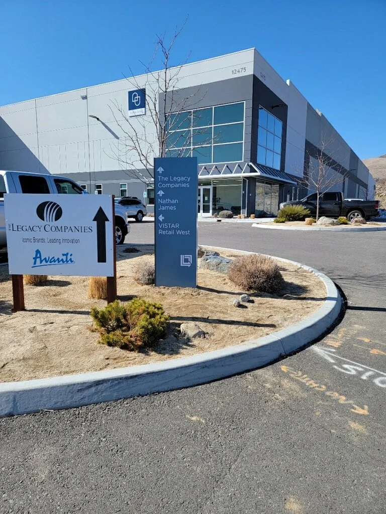 Modern office building with signs indicating directions to The Legacy Companies, Nathan Names, and VISTAR Retail West, with a parking lot and sparse landscaping in the foreground.