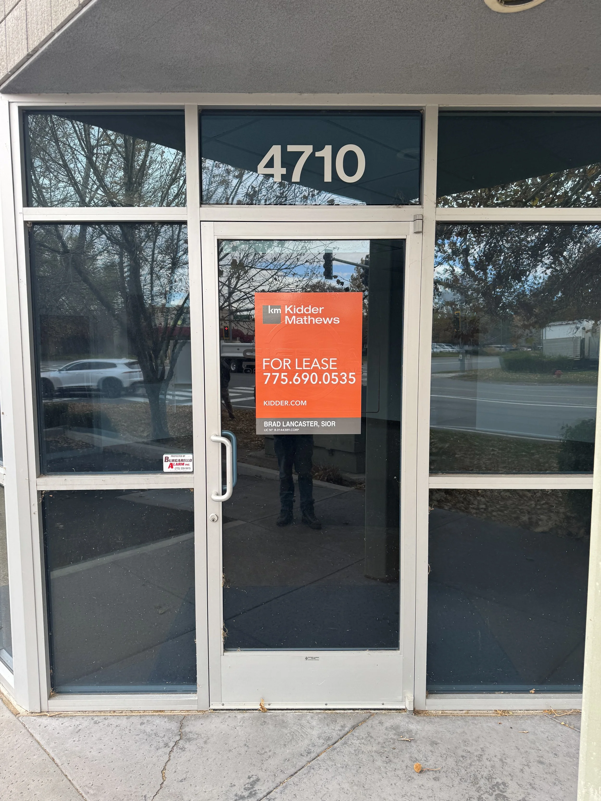 Glass storefront with the number 4710 on the top panel, a bright orange sign in the window advertising a space for lease with contact information, and a reflection of trees, cars, and a person taking the photo.
