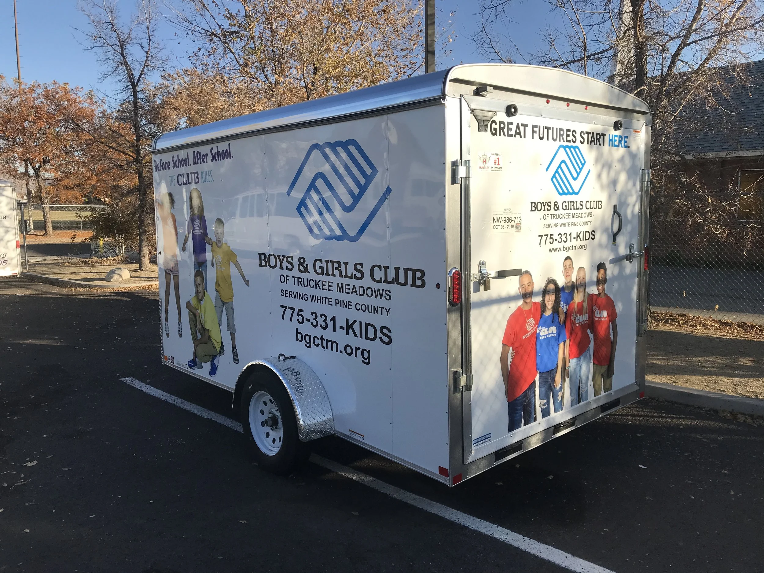 A trailer promoting the Boys & Girls Club of Truckee Meadows, serving White Pine County. The trailer features images of children and adults, along with the organization's logo and contact information, parked on a paved lot with trees and a building i