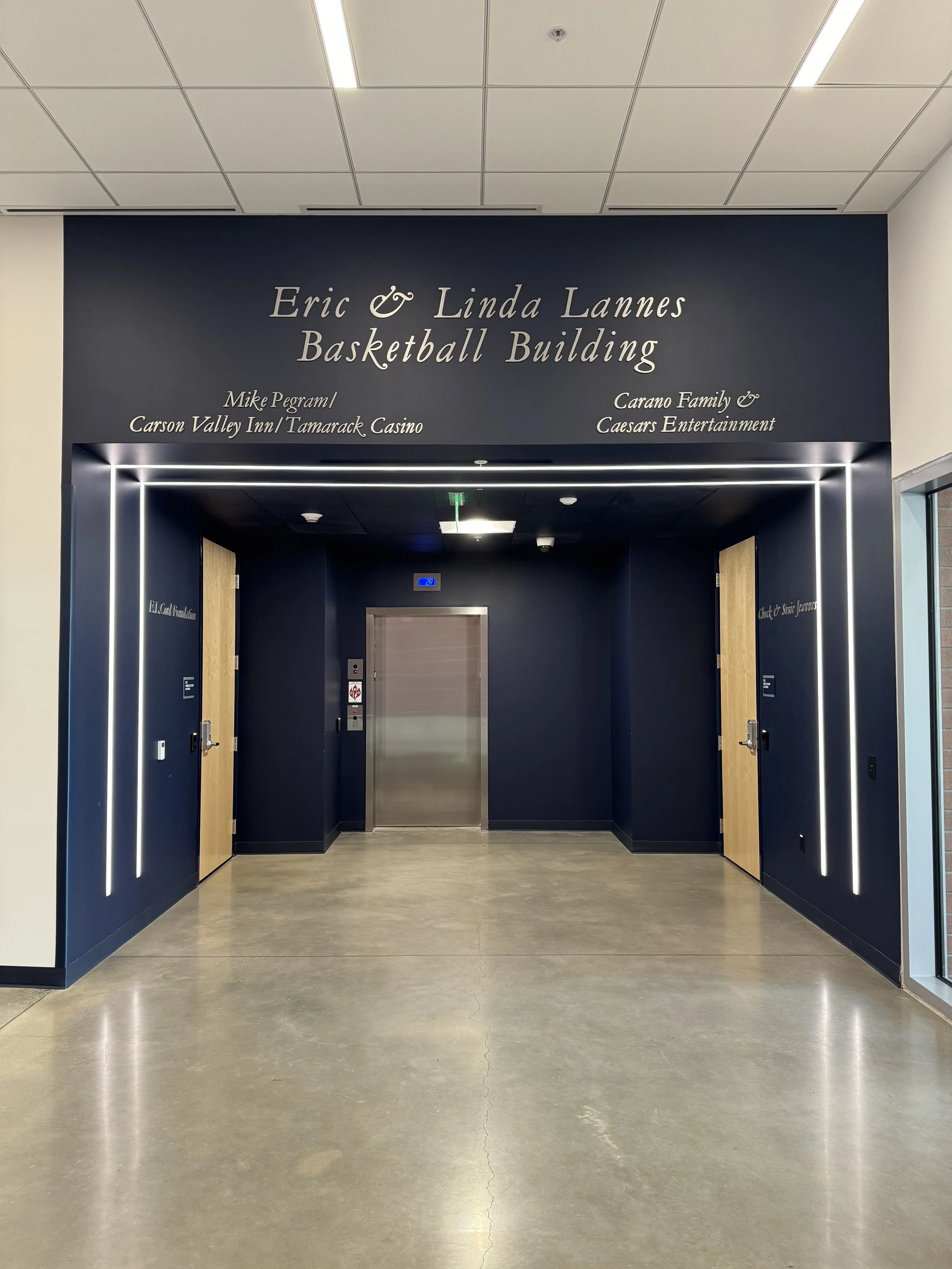 Entrance to the Eric & Linda Lannes Basketball Building with dark blue walls and illuminated door frames, elevator in the center, and plaques on either side of the doors.
