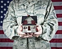 A person in military camouflage holding a small model house in front of an American flag backdrop.
