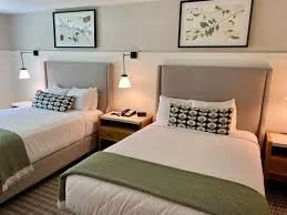 Two beds in a hotel room, each with a pillow and green bed runner, nightstand with a telephone, and artwork on the wall above.