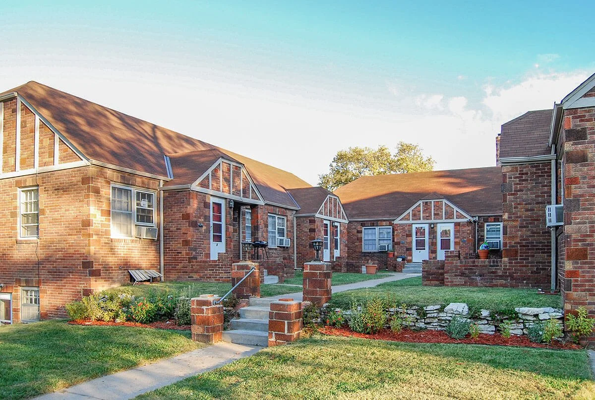 A courtyard with several brick townhouse buildings featuring white trim, small front steps, potted plants, garden beds, and a concrete path, under a blue sky with scattered clouds.