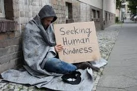 Person sitting on a sidewalk holding a sign that reads 'Seeking Human Kindness'.