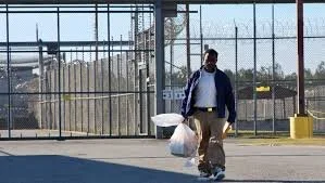 Man walking outdoors near a metal fence, carrying shopping bags and a cardboard box.
