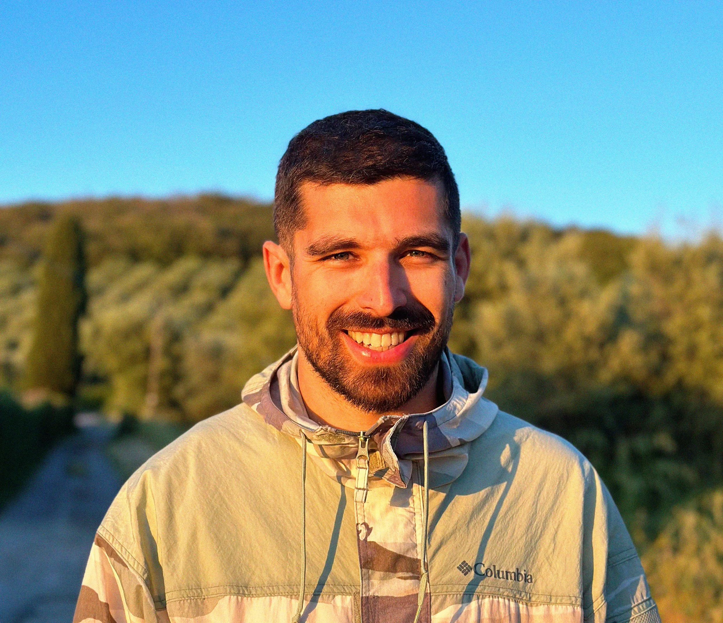 Smiling man with short dark hair and beard outdoors during sunset, wearing a beige Columbia jacket, with trees and blue sky in the background.