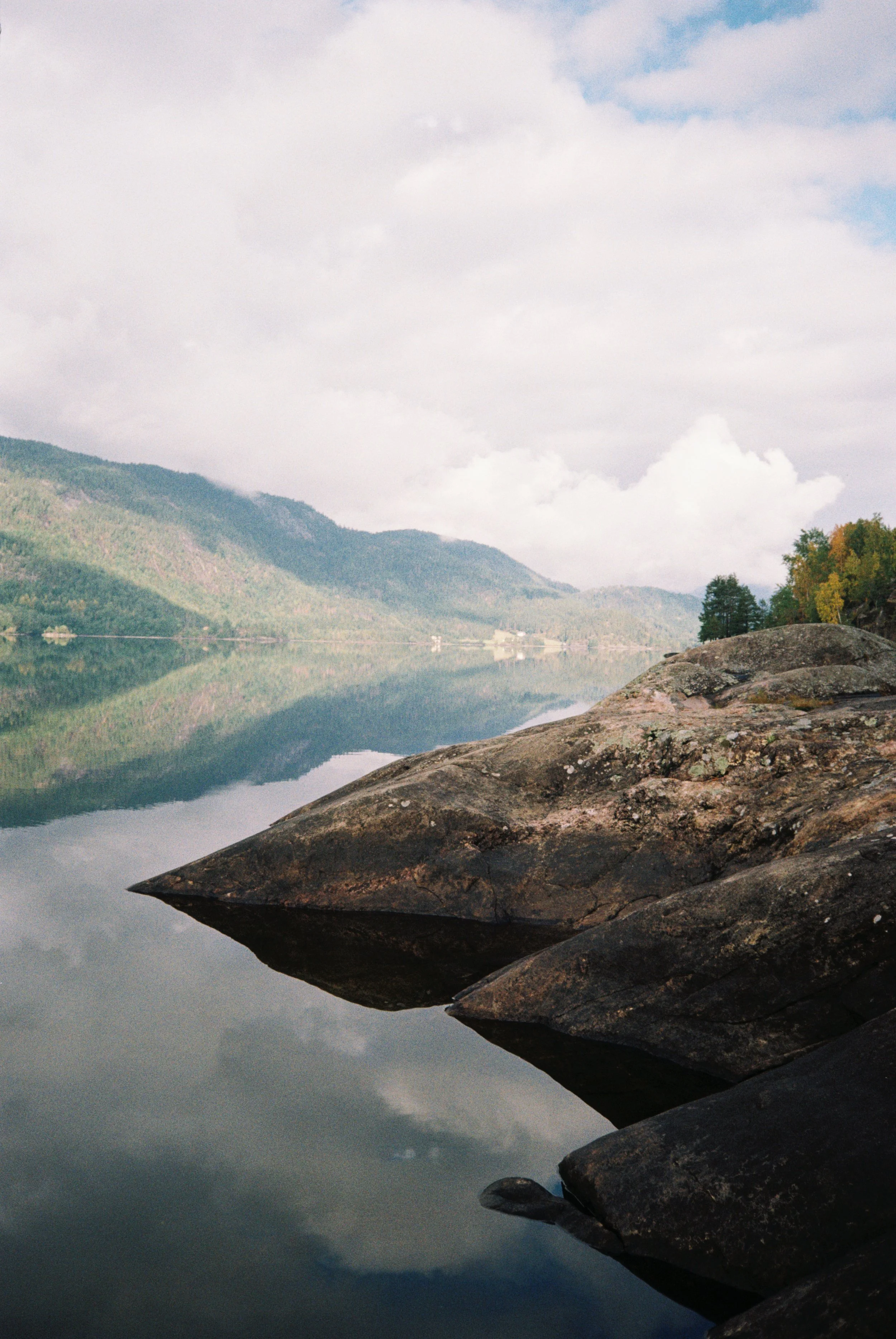 Mountains and trees reflected on a calm lake with rocks in the foreground and a partly cloudy sky.