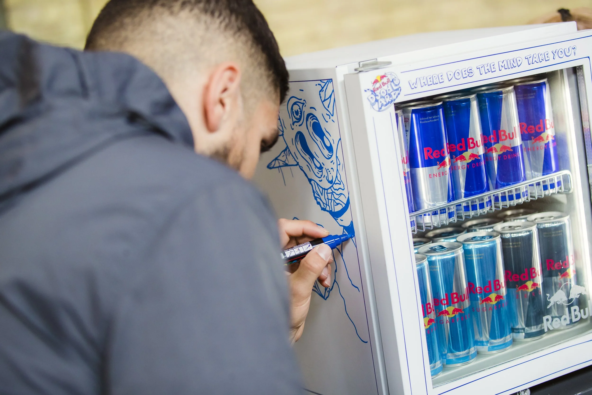 A man with short dark hair and a beard is drawing a blue line on a white backdrop with a marker. The backdrop has an illustration of a creature's face. Nearby, a mini fridge with a glass door contains cans and bottles of Red Bull energy drinks.