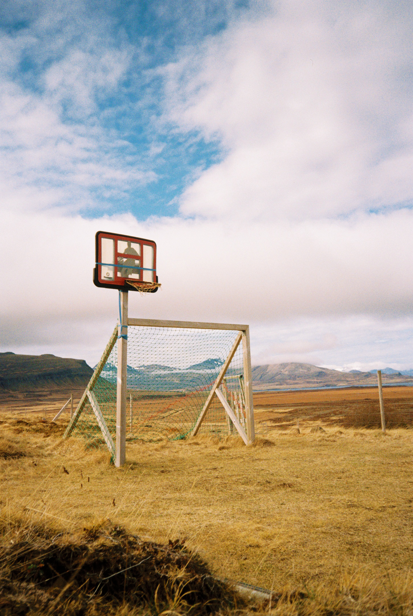 An outdoor basketball hoop with a backboard and a net, placed over a soccer goal on a dry, grassy field with mountains in the background under a partly cloudy sky.