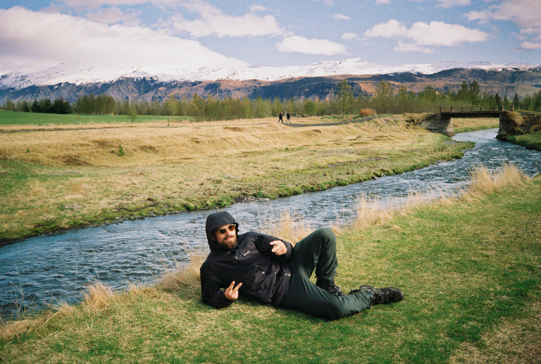 A man with dark hair, beard, sunglasses, wearing a black hoodie and pants, lying on the grass beside a small river in a scenic landscape with snow-capped mountains in the background, pointing towards the camera.