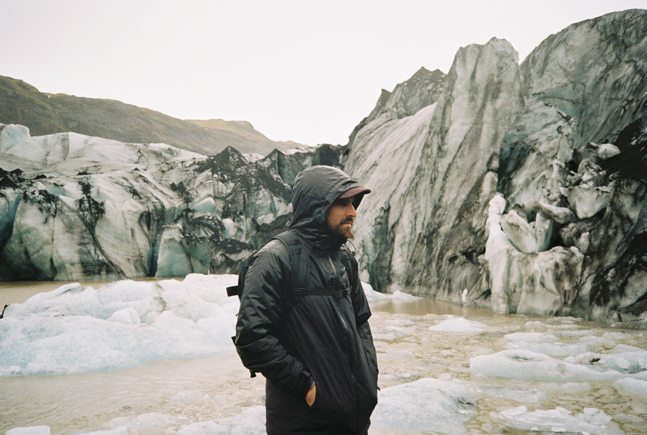 A man in a dark rain jacket with a hood and backpack standing by icy water with a large glacier and mountains in the background.