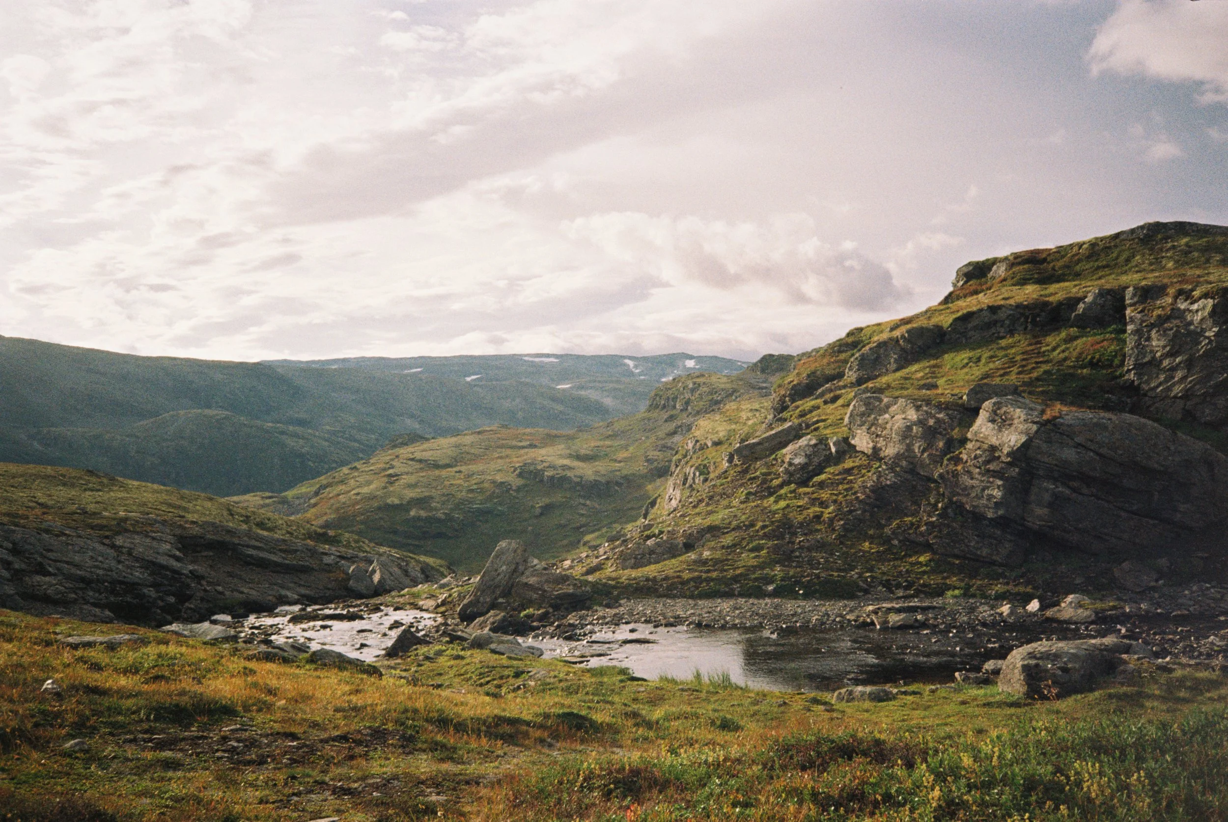 A scenic view of a mountain valley with a small river running through grassy terrain. Large rocks and hillsides covered in green vegetation are visible, with a cloudy sky overhead.